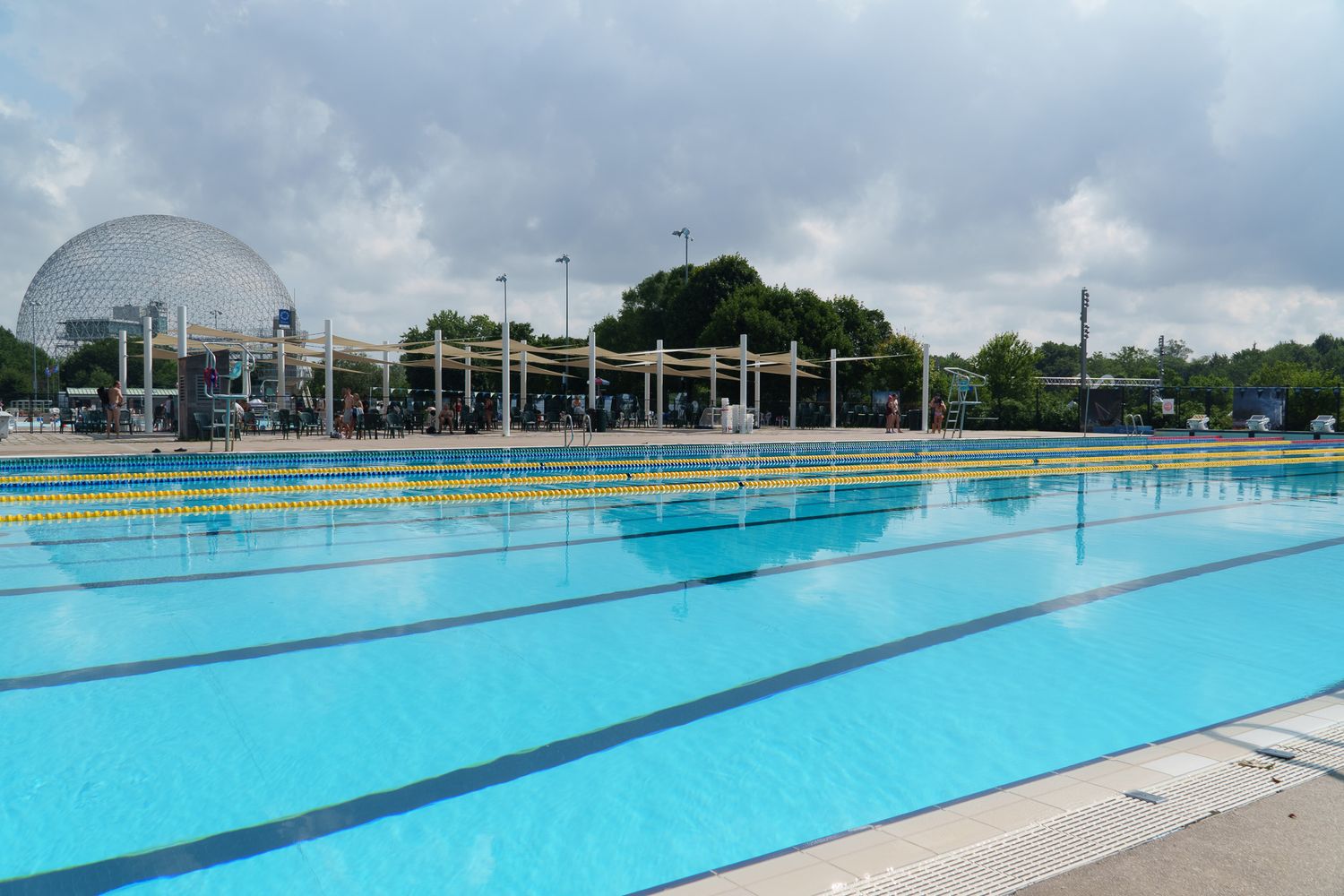 Outdoor competition swimming pool with blue lanes and a large geodesic dome structure visible in the background under cloudy skies.