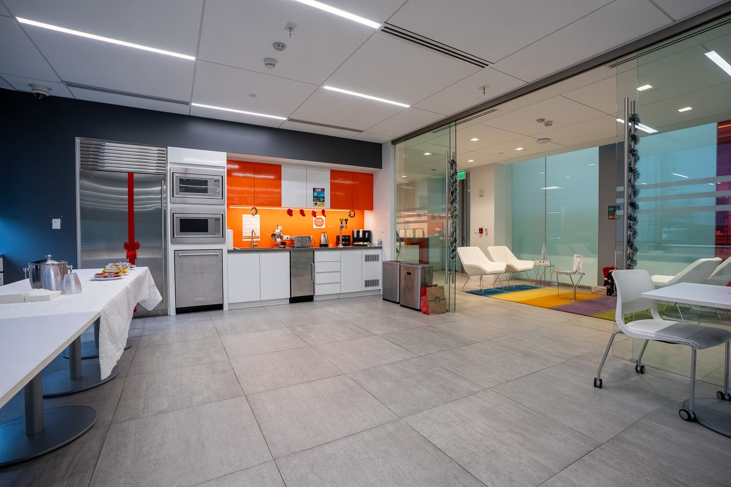 Modern office break room with orange kitchen cabinets, stainless steel appliances, and glass partition walls.