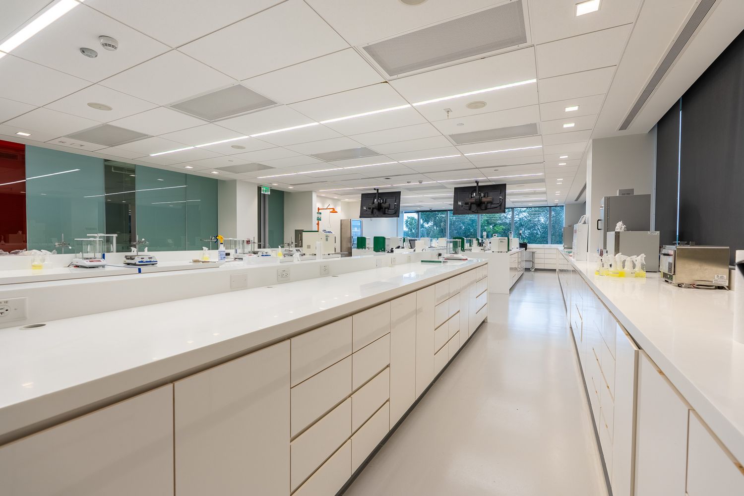 Modern laboratory interior with white countertops, cabinets, and equipment under bright fluorescent lighting.