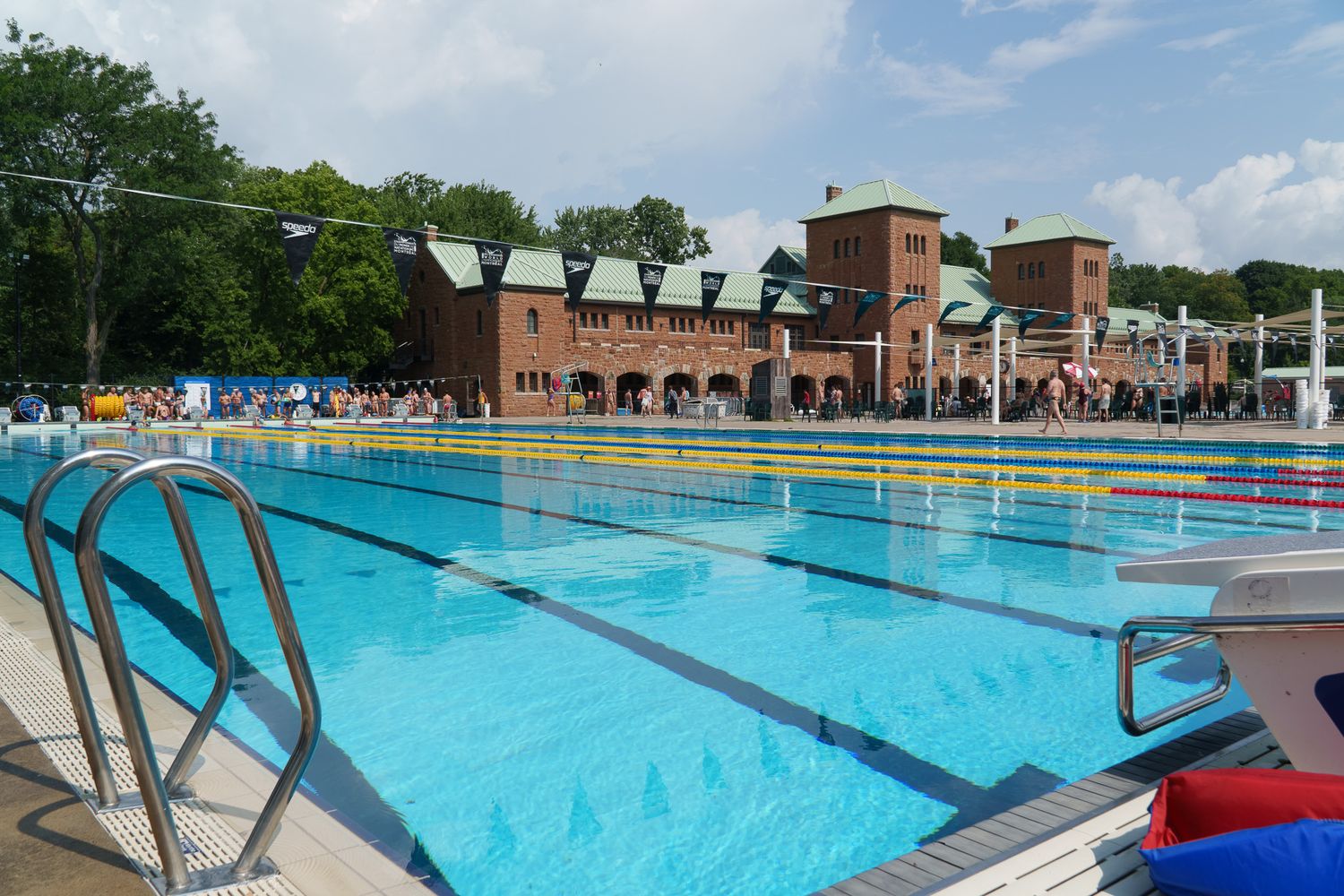 Outdoor Olympic swimming pool with clear blue water, metal railings, and a historic brick building surrounded by trees.