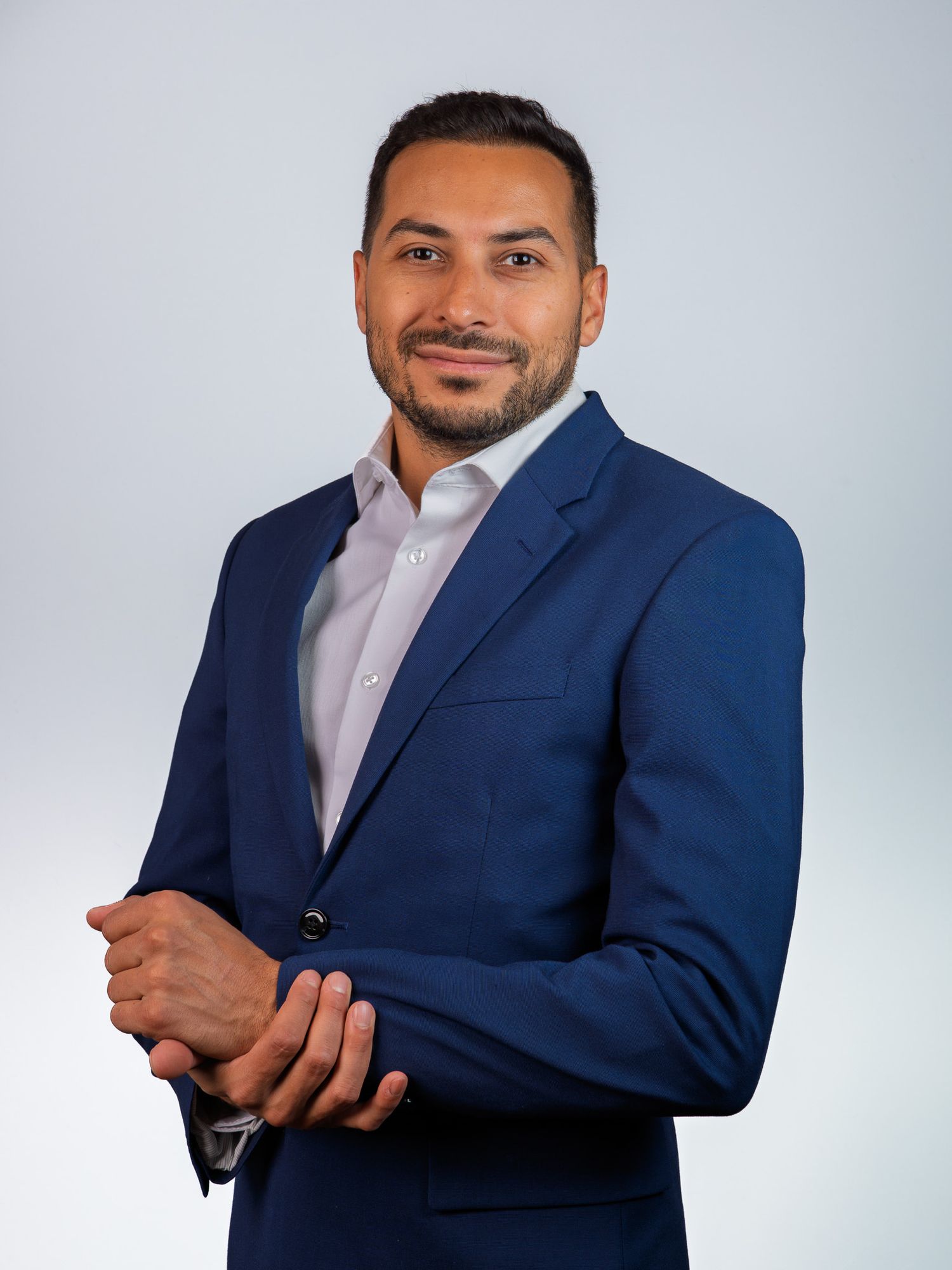 Professional man in navy blue suit with white shirt, arms crossed, posing confidently against light gray background.