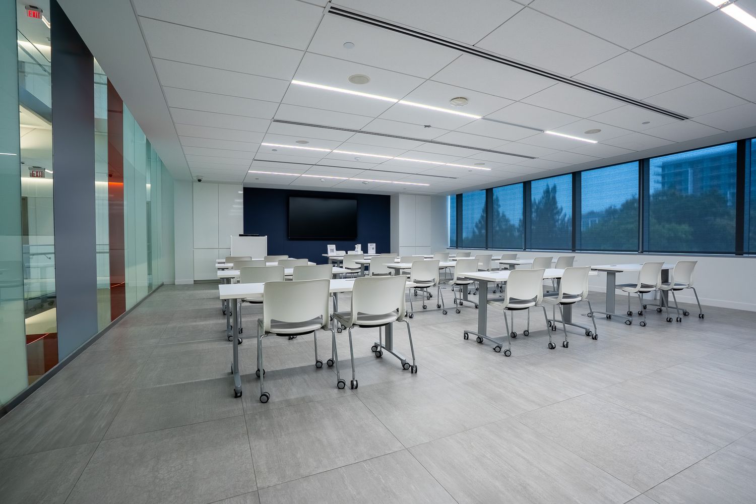 Modern conference room with white chairs, folding tables, large display screen, and floor-to-ceiling windows in a bright office.