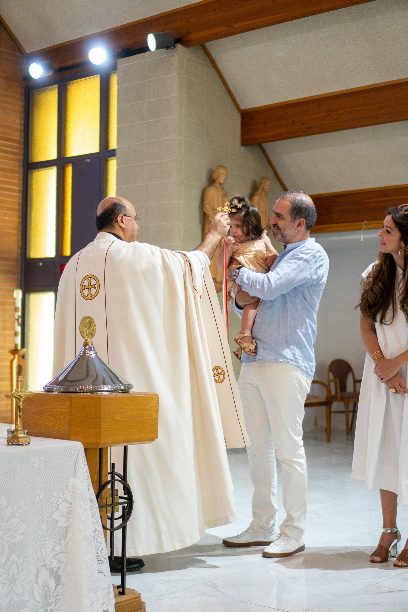 Priest performing a baptism ceremony, anointing a baby held by father in a modern church.