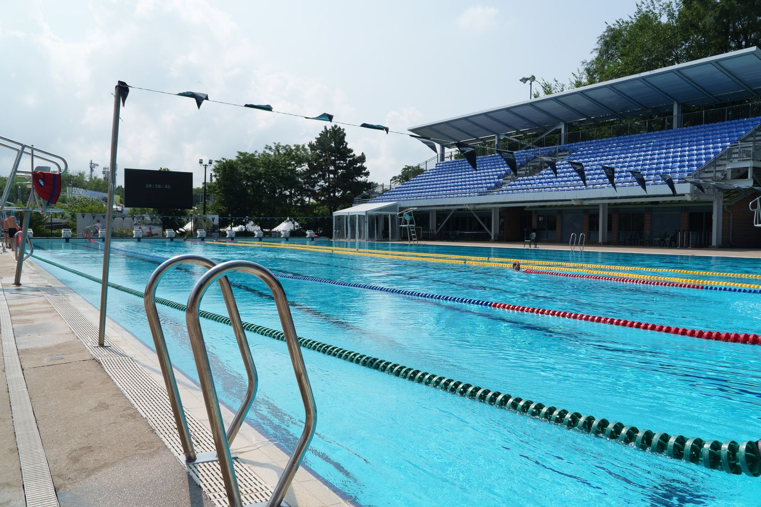 Outdoor Olympic swimming pool with blue lane dividers, metal railings, and a blue stadium grandstand under sunny skies.