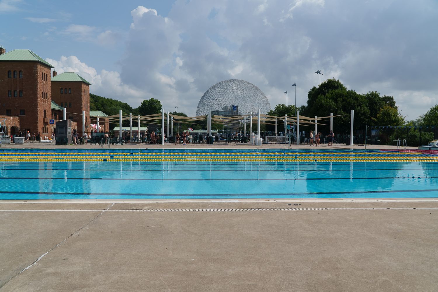 Outdoor Olympic swimming pool with clear blue water, a geodesic dome structure in the background under a partly cloudy sky.
