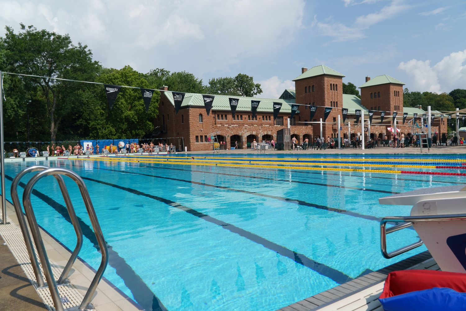 Outdoor Olympic swimming pool with clear blue water, lane dividers, and a red brick building surrounded by trees in the background.