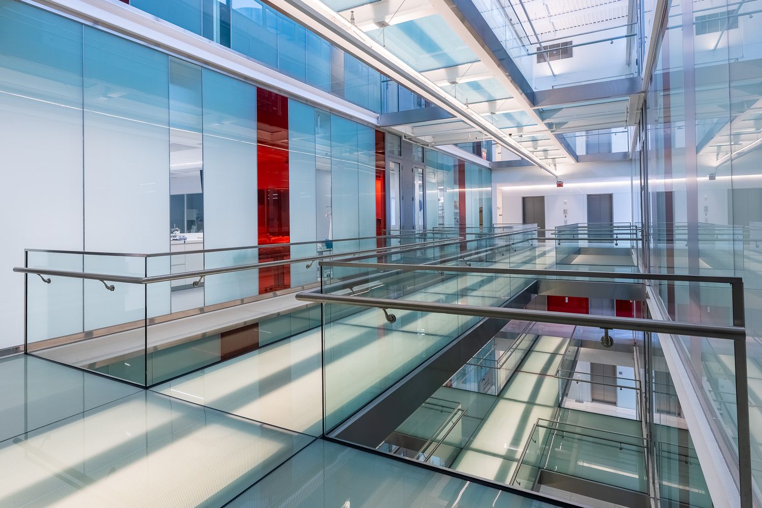 Modern glass atrium interior with transparent floors, steel railings, and vibrant red accents in a contemporary office building.