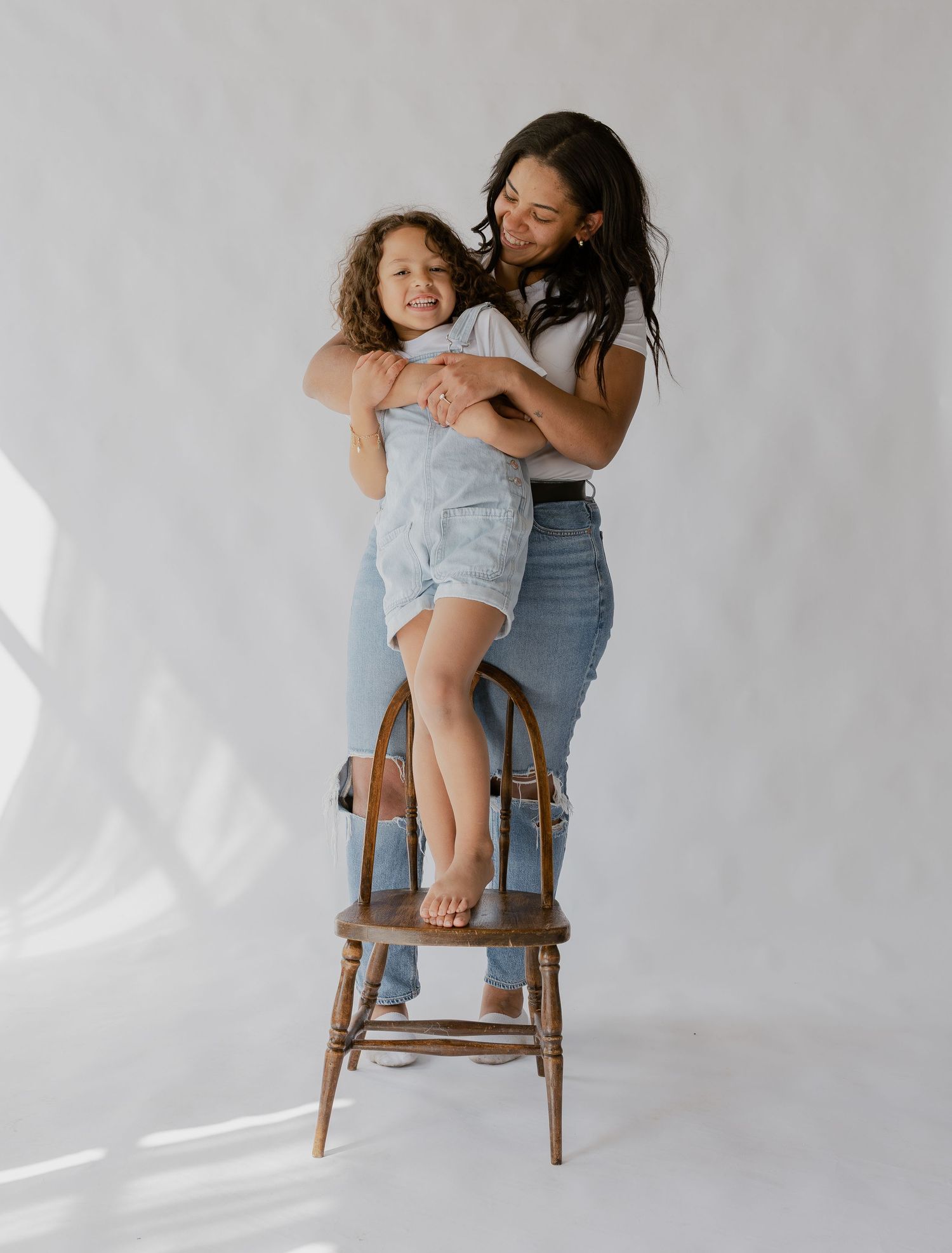 A woman hugs a smiling child standing on a wooden chair in a bright white studio setting.