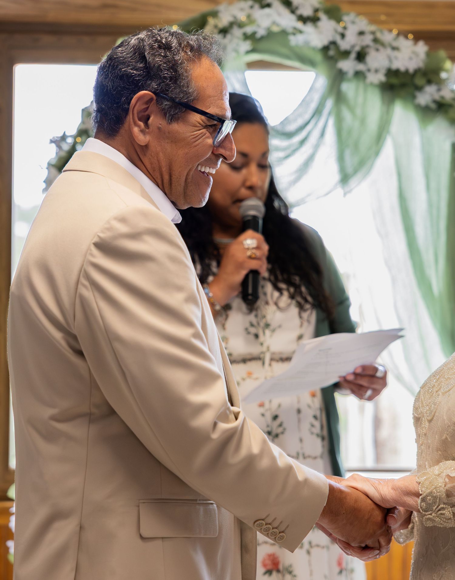 Man in beige suit reading vows at wedding ceremony with officiant holding microphone in background.
