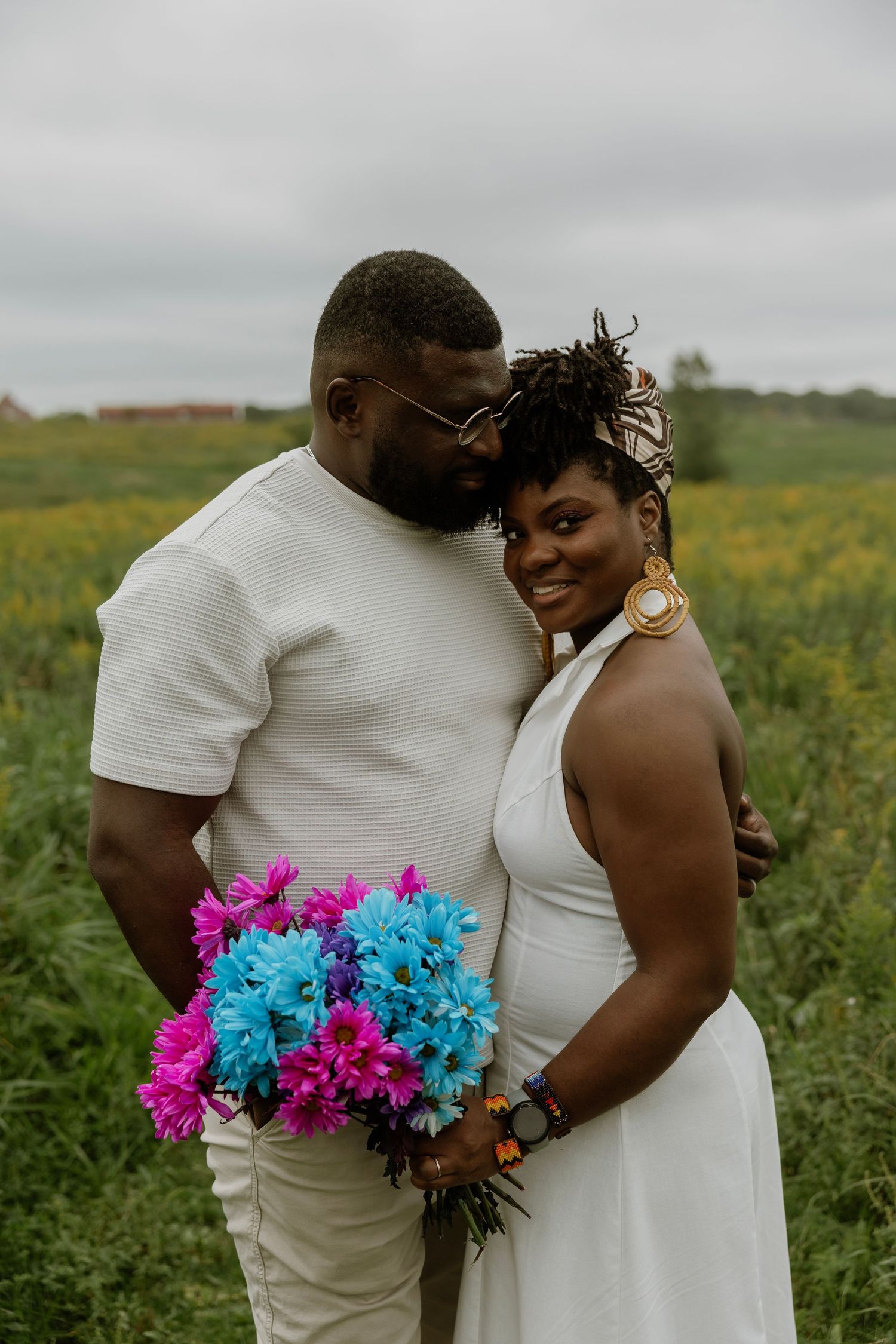 Couple in white attire poses in a flower field, holding vibrant pink and blue bouquet, celebrating their wedding day.