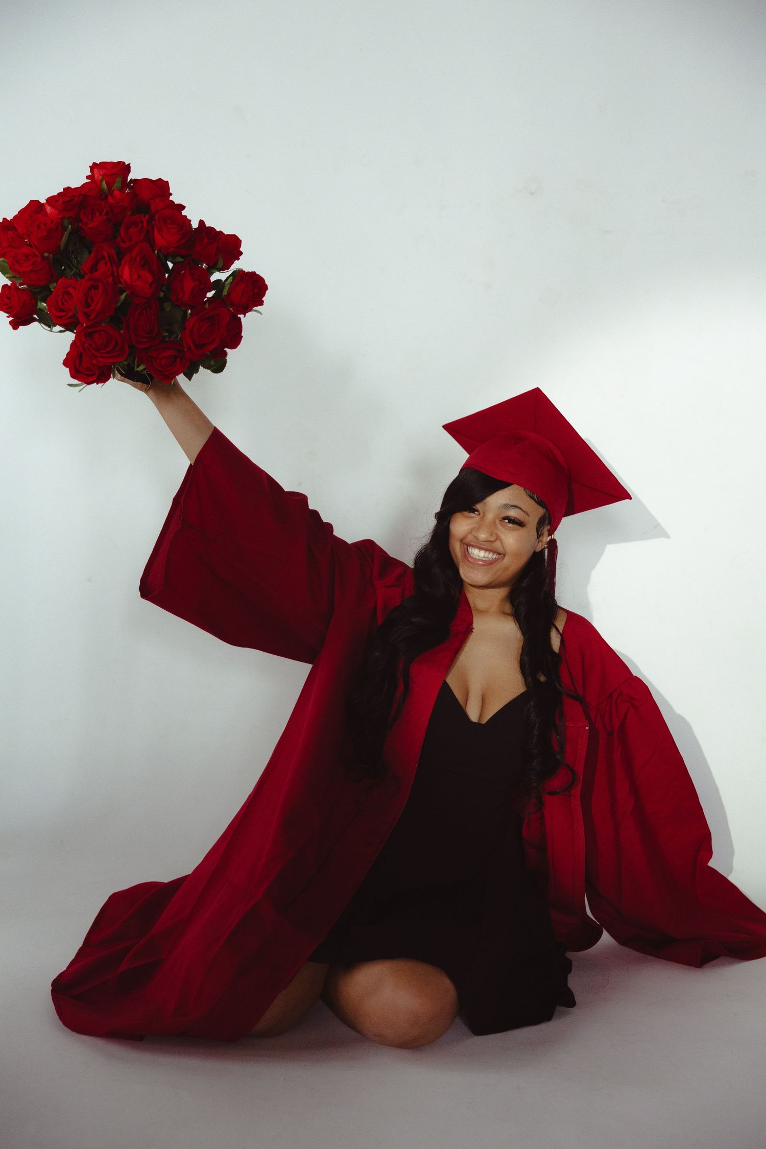 Smiling graduate in red cap and gown sitting on floor holding bouquet of red roses in celebratory pose.