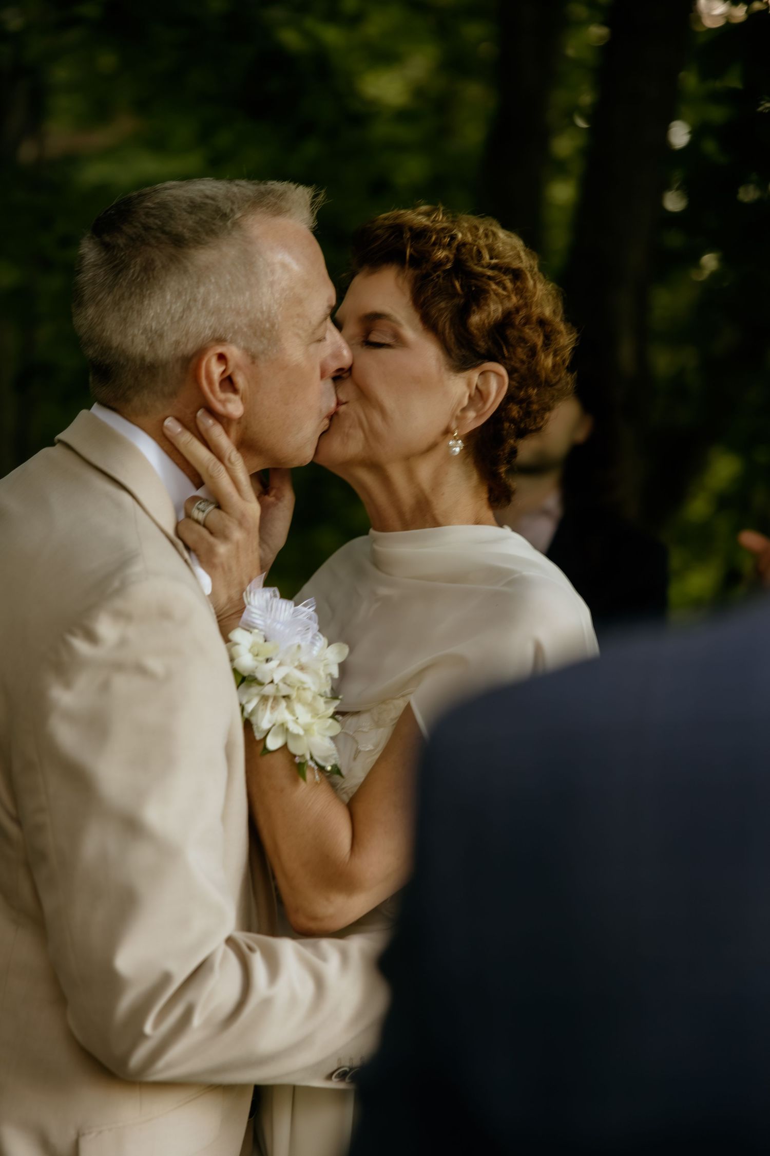 Mature couple sharing a first kiss at outdoor wedding ceremony, groom in beige suit, bride in white dress with floral corsage.