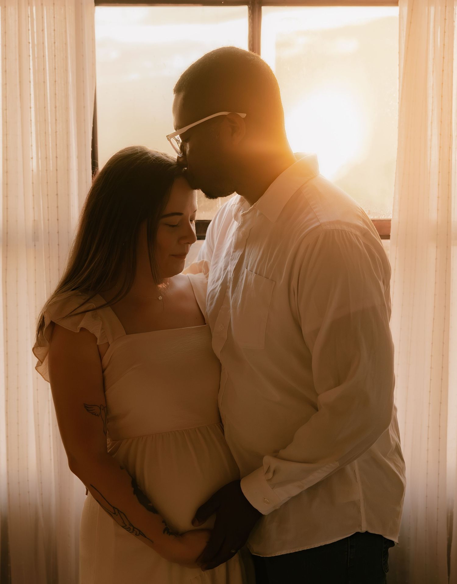 A couple shares a tender moment bathed in golden sunset light by a window, the woman in a white maternity dress.