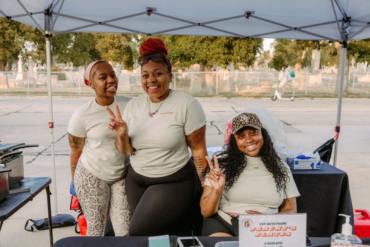 Three women smiling and posing with peace signs at an outdoor community event booth under a canopy.
