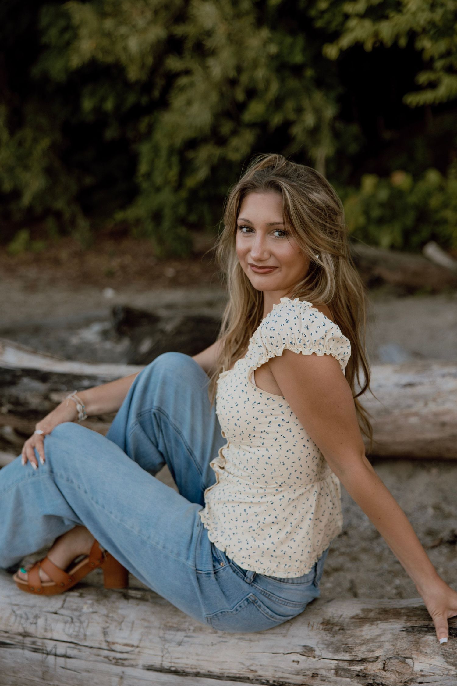 Young woman in white top and jeans sitting on driftwood logs with lush green trees in background.