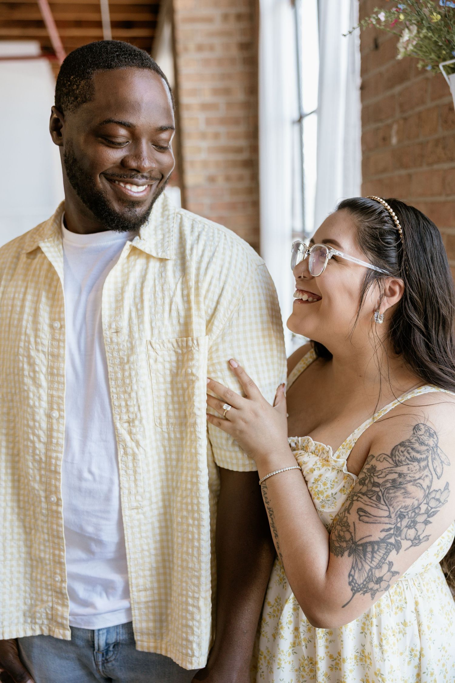 A smiling interracial couple in casual summer outfits, woman with tattoos touching man's shirt indoors.