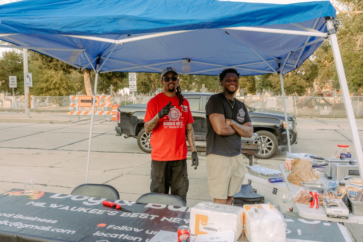 Two men smiling under a blue canopy tent at an outdoor BBQ food vendor booth with supplies on the table.