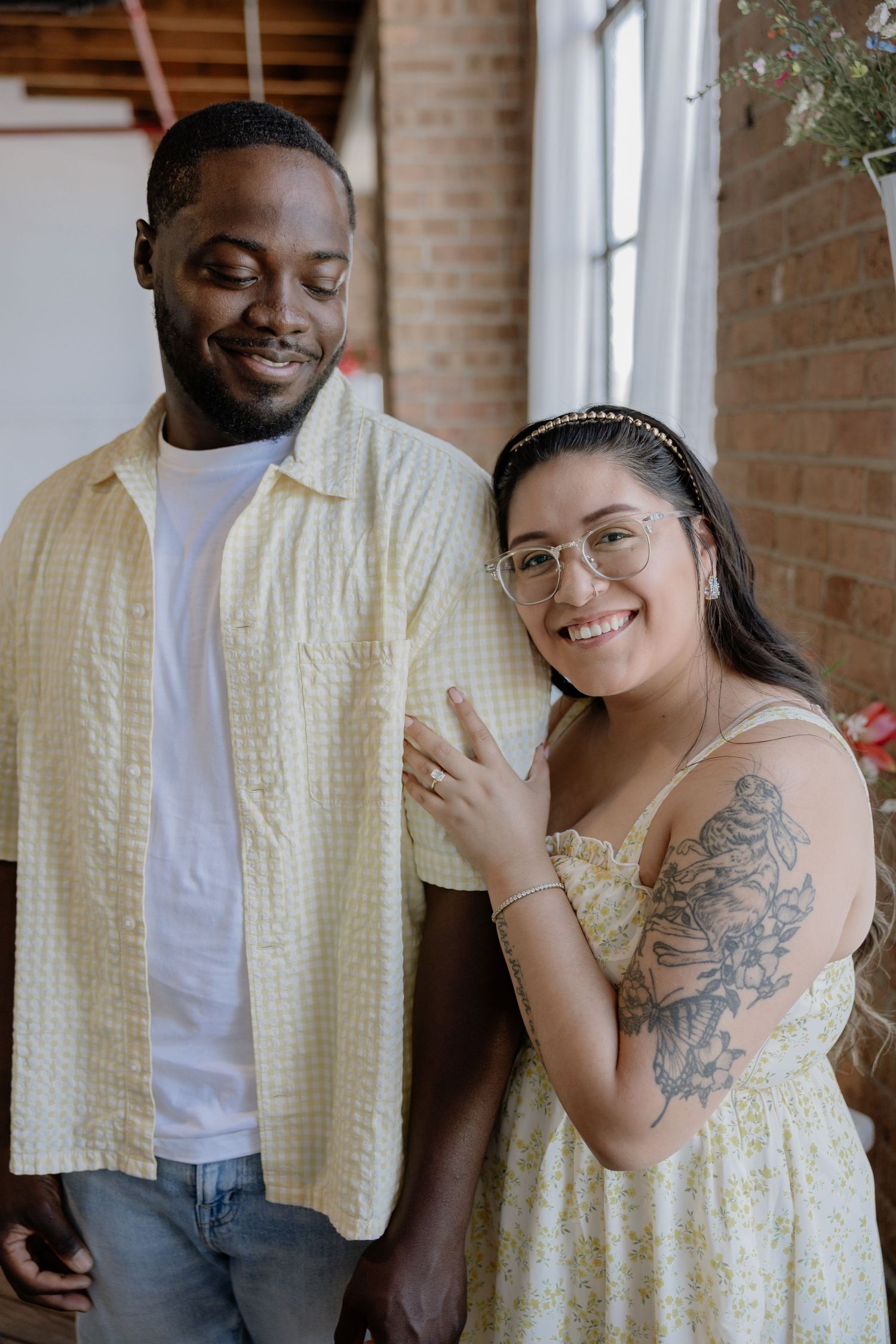 A smiling couple poses indoors, the woman wearing glasses and a floral tattoo, both dressed in yellow outfits.