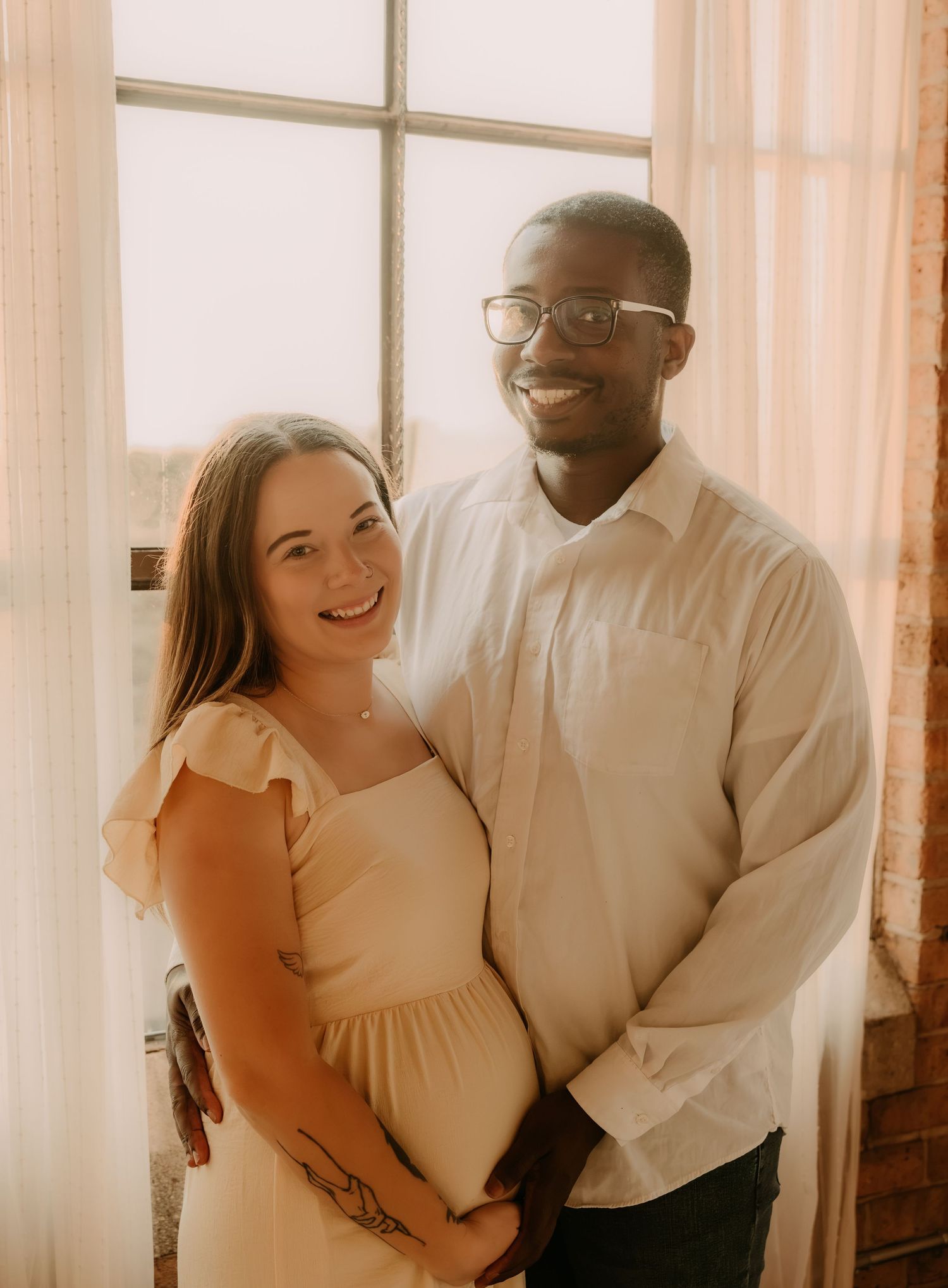 Happy interracial couple posing together near a bright window, woman in cream dress showing baby bump.
