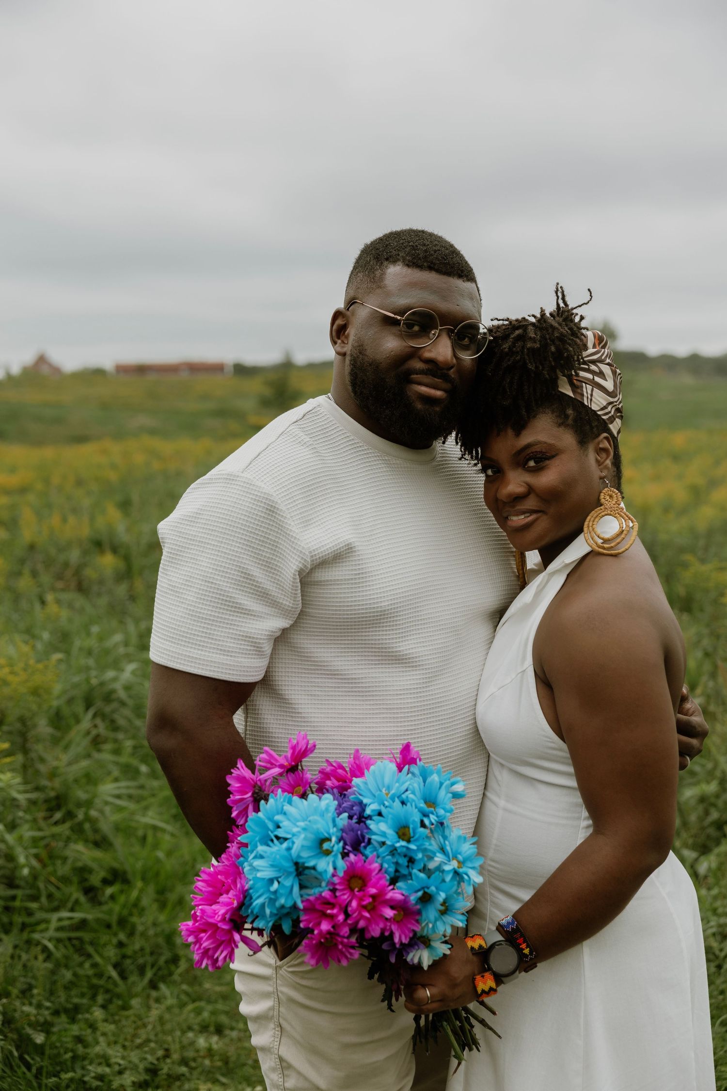 Couple in white attire poses in a flower field, woman holding vibrant pink and blue bouquet, smiling outdoors.