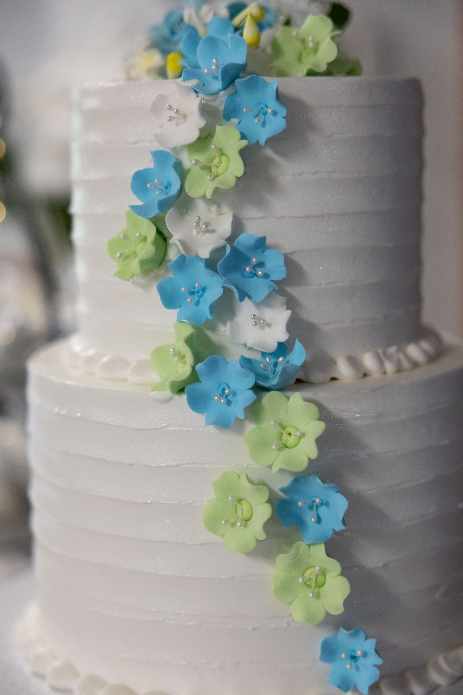 Three-tier white wedding cake decorated with cascading blue, green, and white sugar flowers on textured frosting.