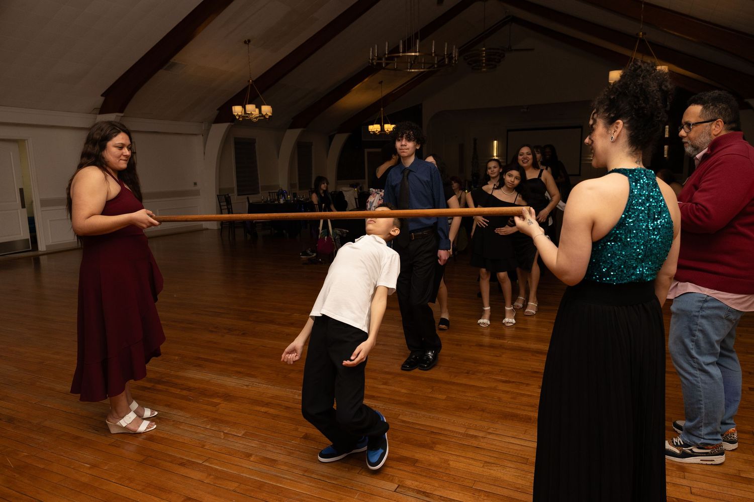 People playing limbo at a formal dance party in a wood-floored ballroom, with a boy bending under a ribbon.
