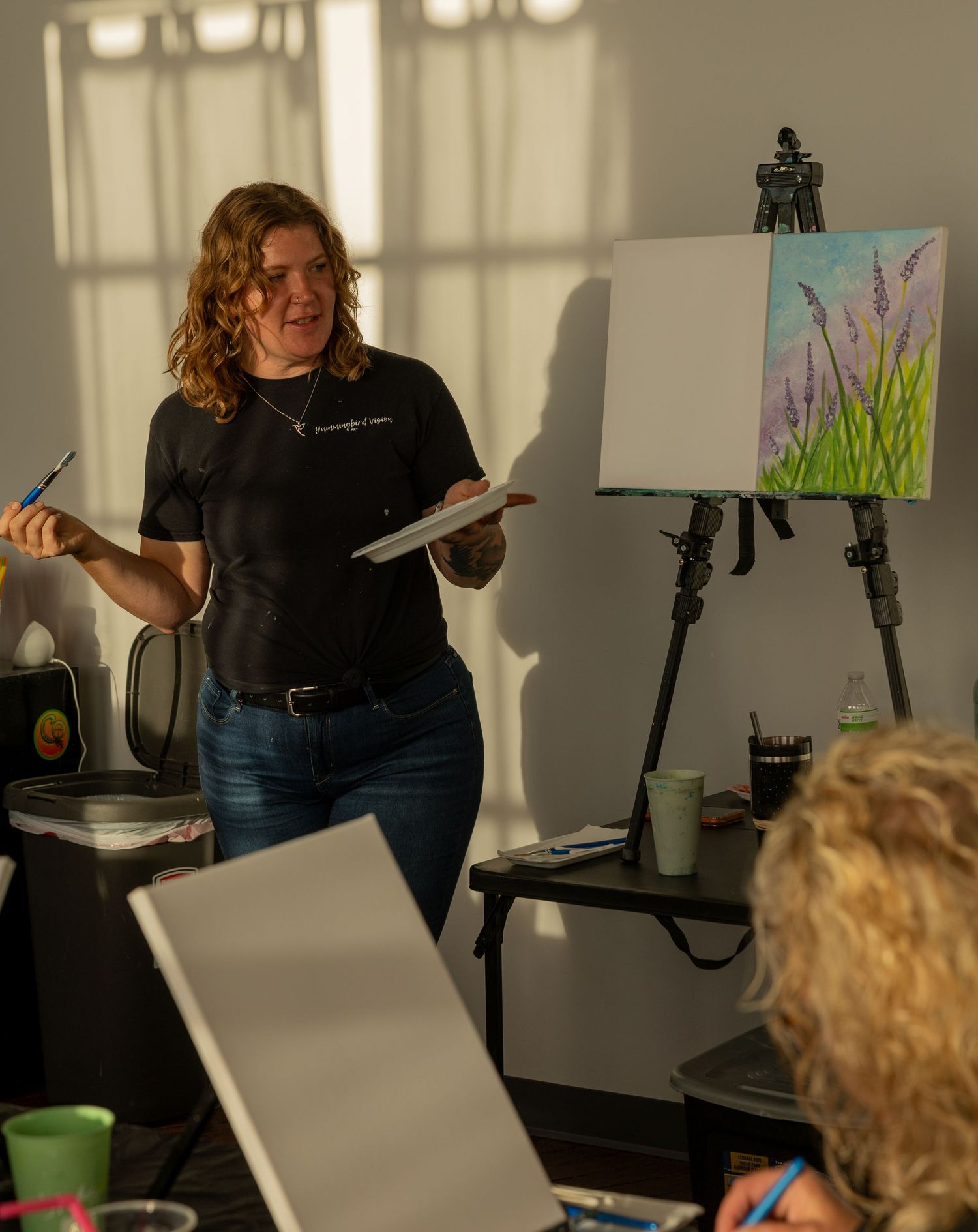 Art instructor holding paper and paintbrush while teaching a painting class with a floral canvas on an easel.