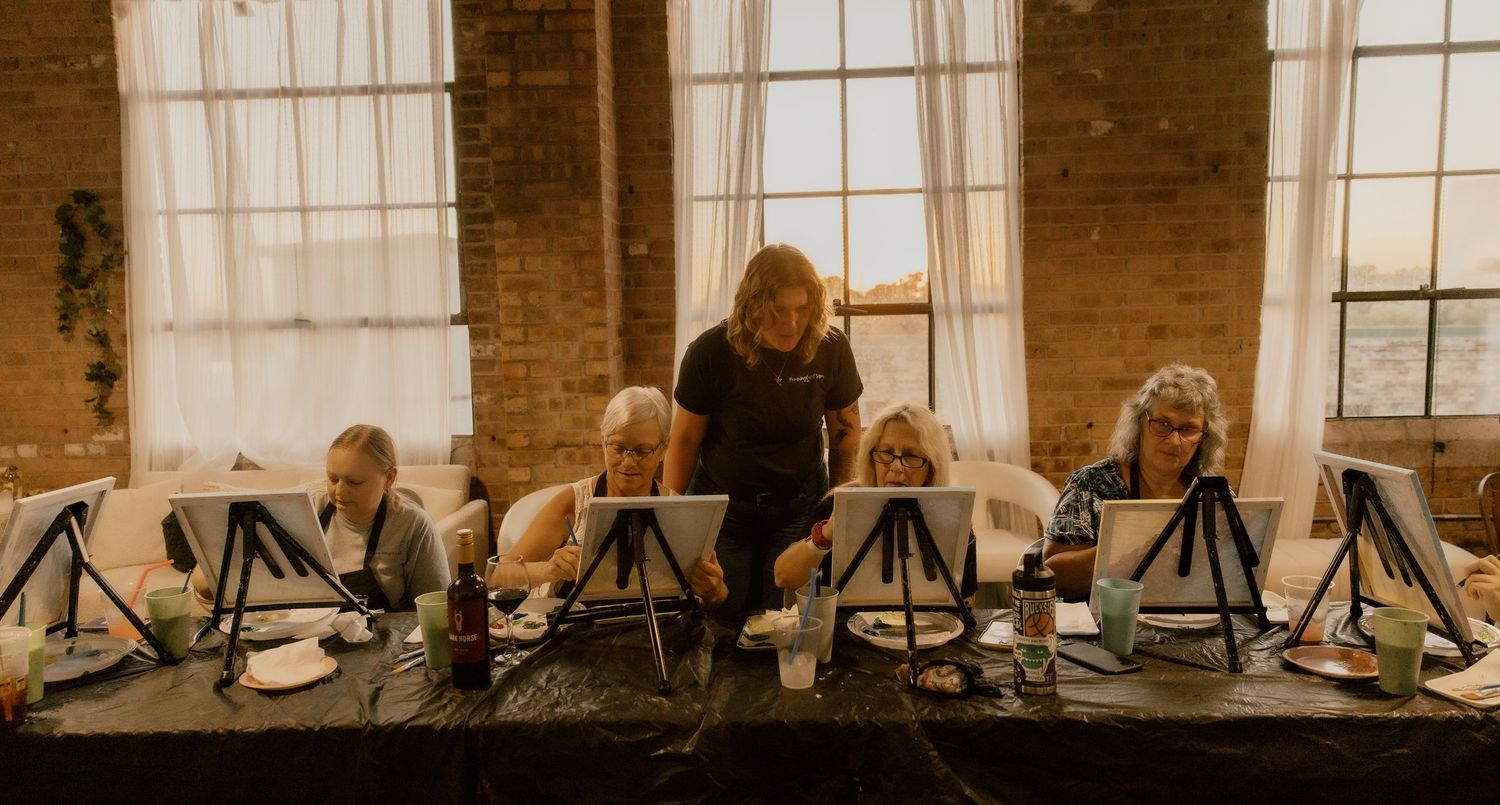 Art instructor guides students painting on easels in a rustic brick-walled studio with large windows.