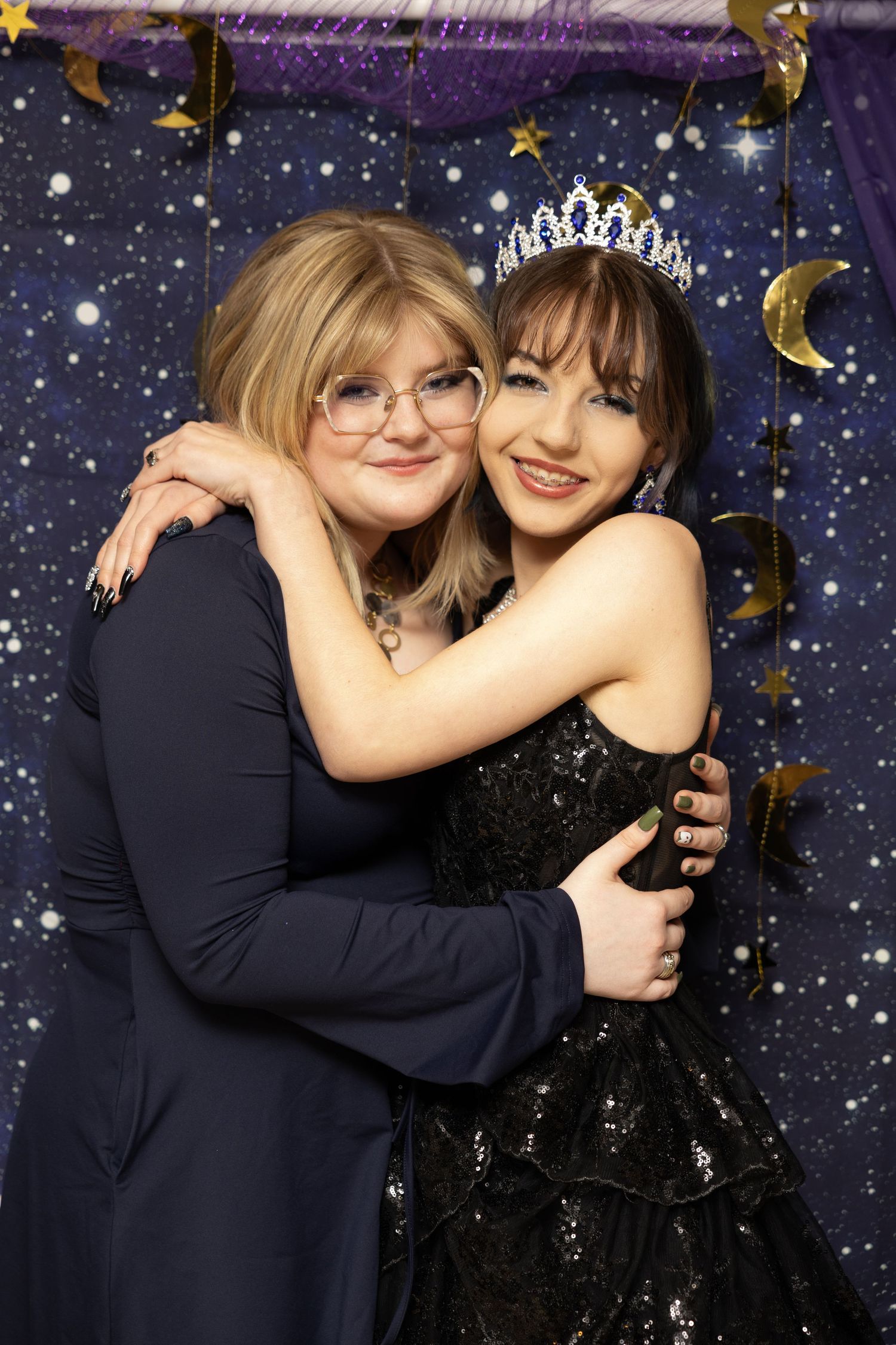 Two young women in formal attire hug and smile at a starry prom photo booth, one wearing a tiara and black gown.