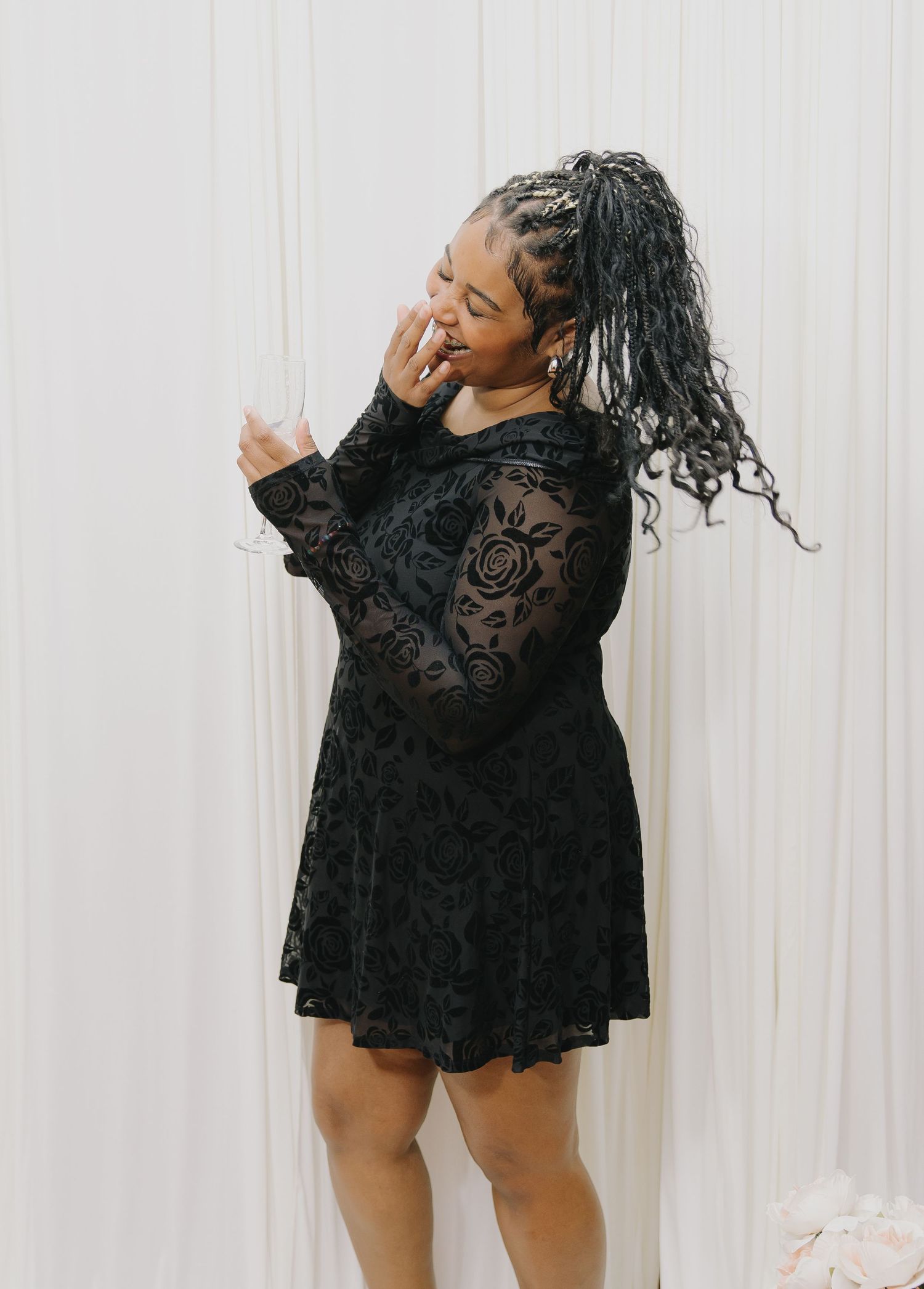 Woman in black floral lace long-sleeve mini dress with braided ponytail, posing gracefully against white curtain backdrop.