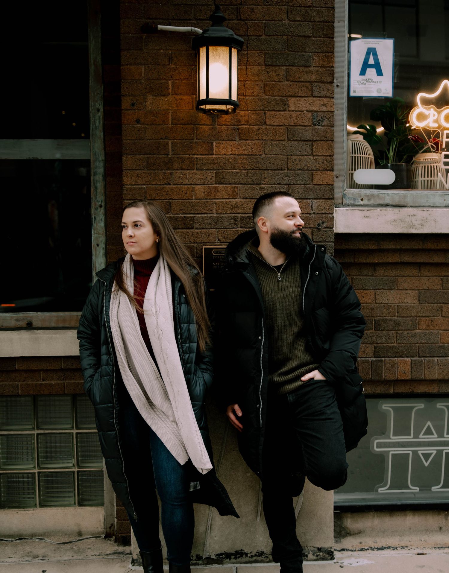 A man and woman in winter coats stand against a brick wall beside a glowing lantern on a city street.
