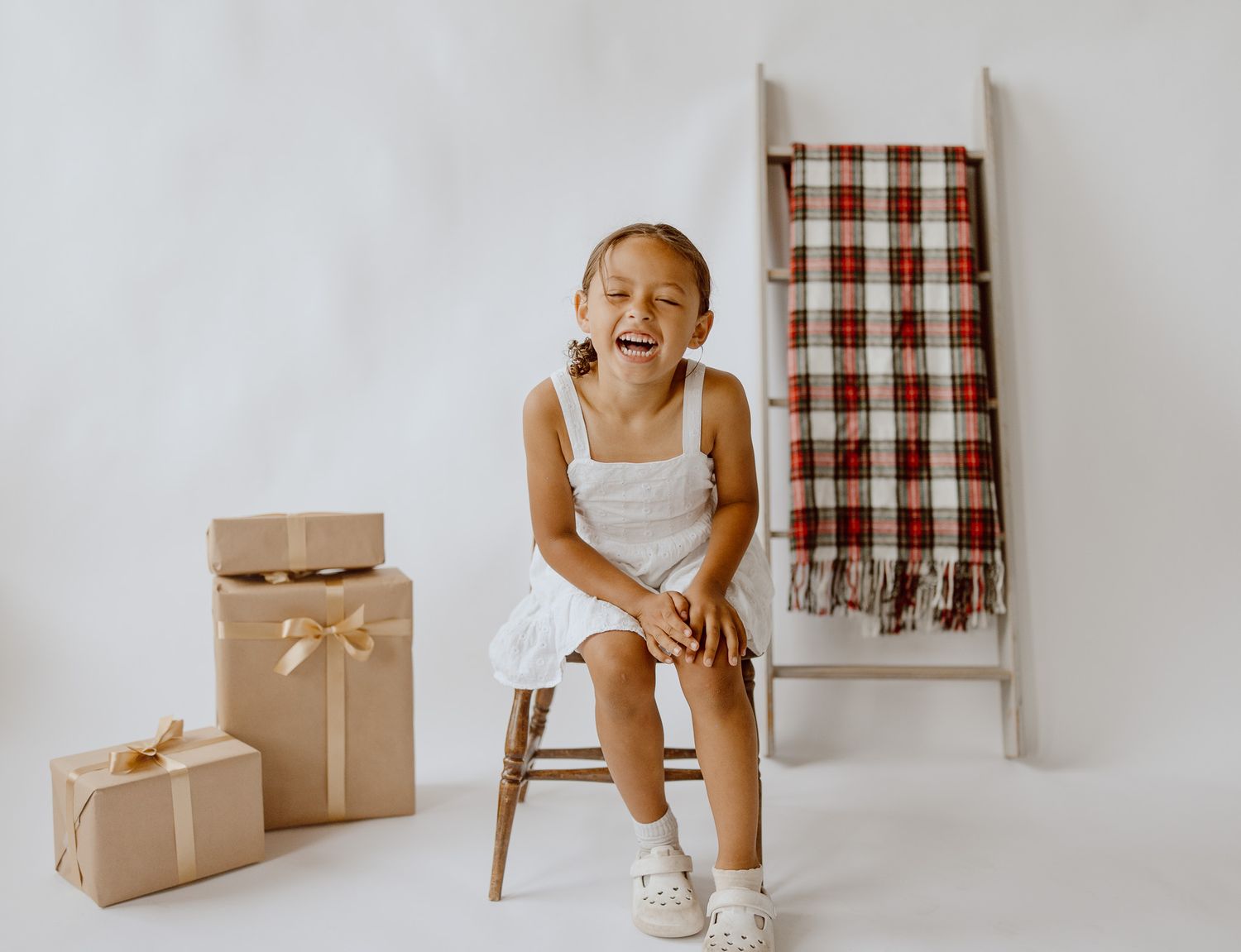 A joyful young girl in a white dress sits on a stool laughing, surrounded by wrapped gifts and a plaid blanket ladder.