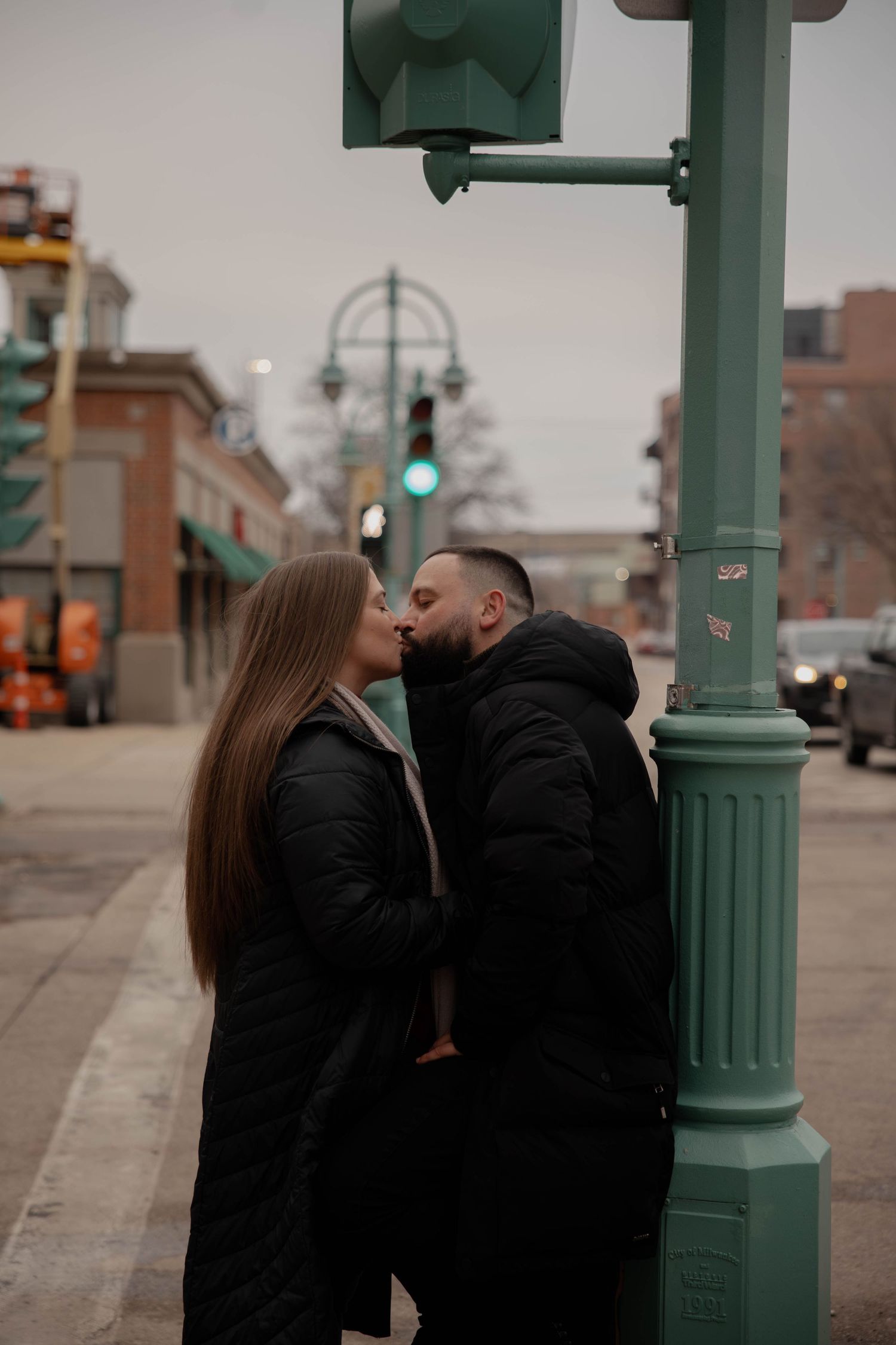 Couple sharing a kiss on a city sidewalk beside a green street lamp post on a cloudy winter day.