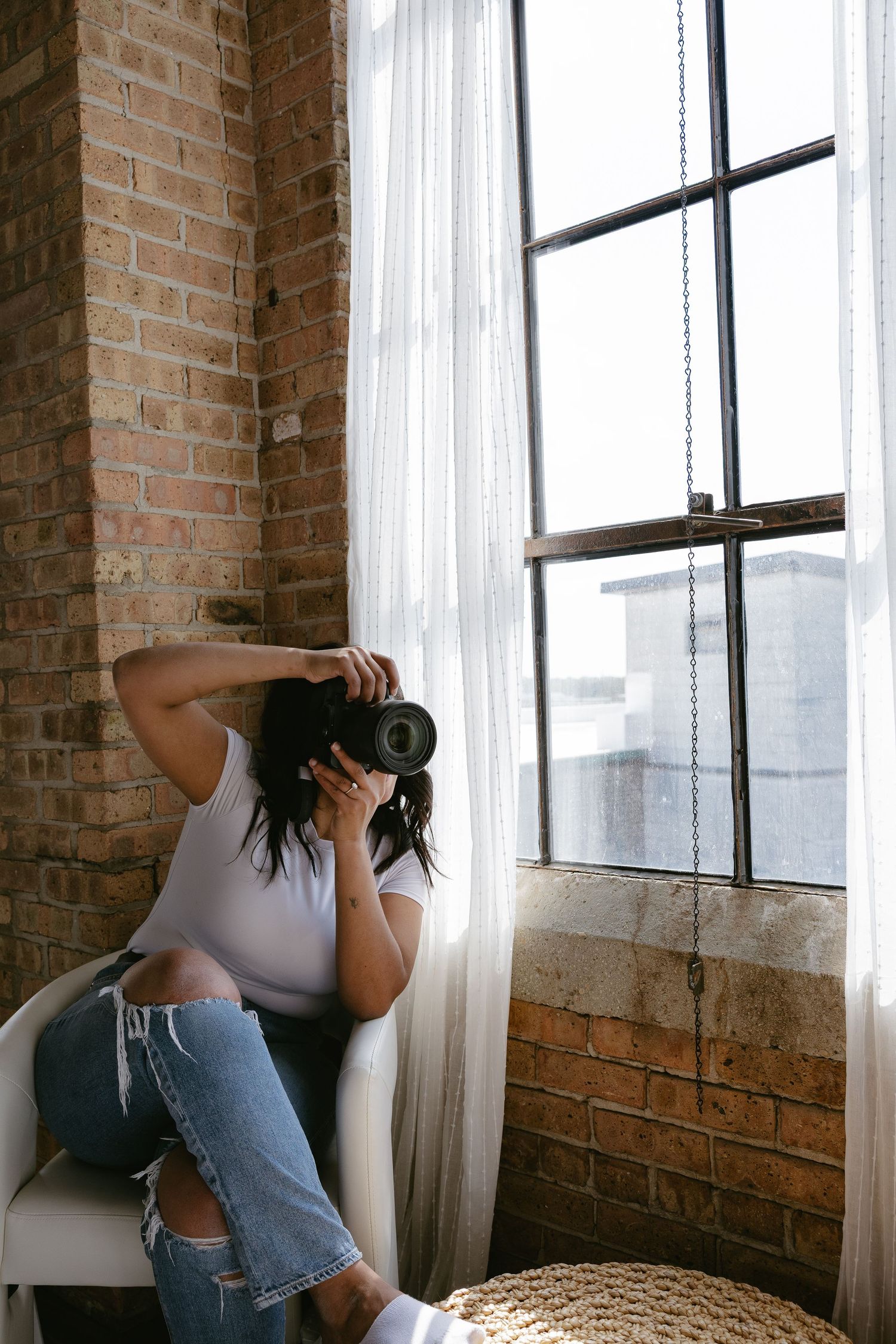 Woman in white tee and ripped jeans sitting in chair by brick wall window, photographing with DSLR camera.
