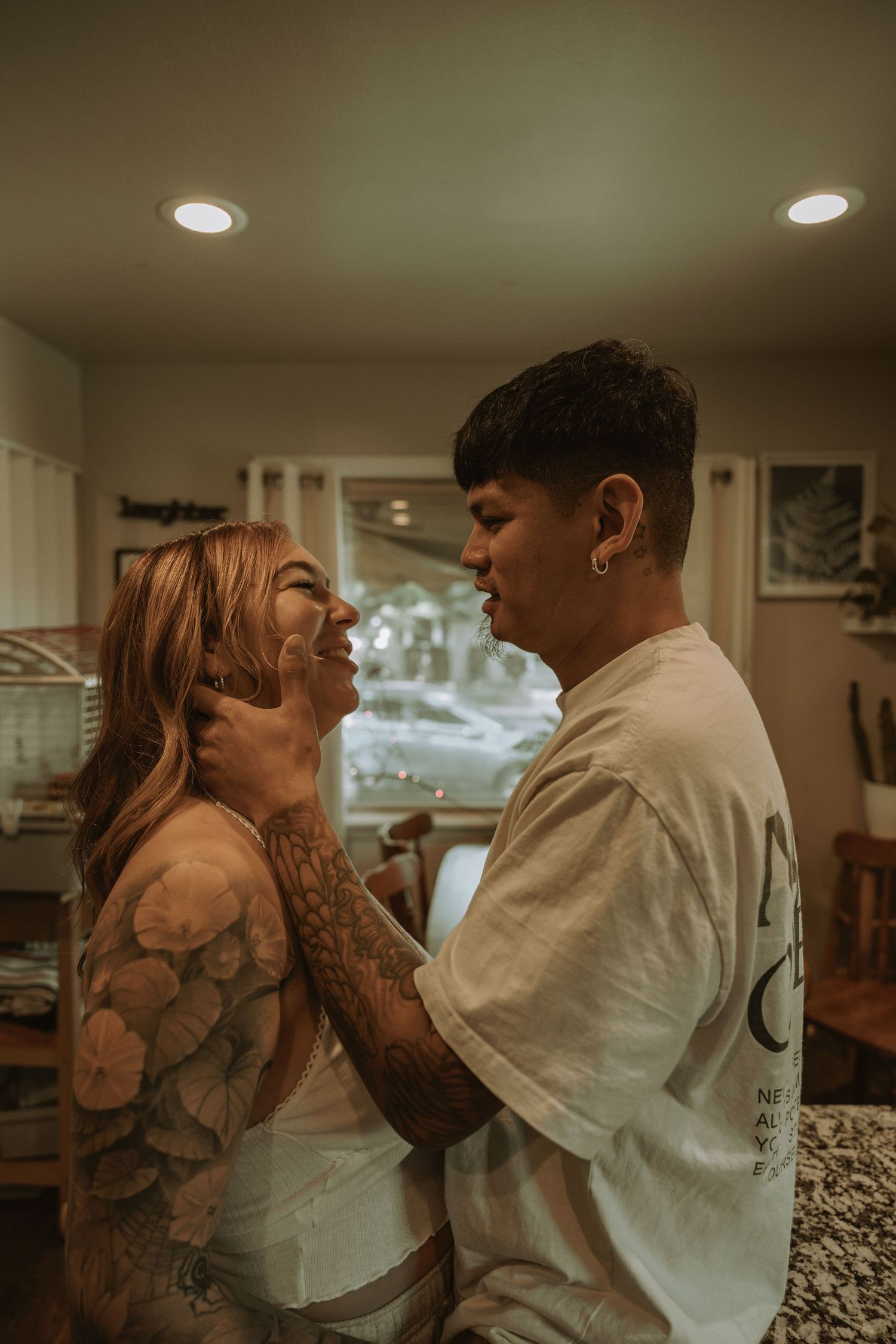 A tattooed couple shares a tender moment indoors, with the man gently holding the woman's face as she smiles up at him.