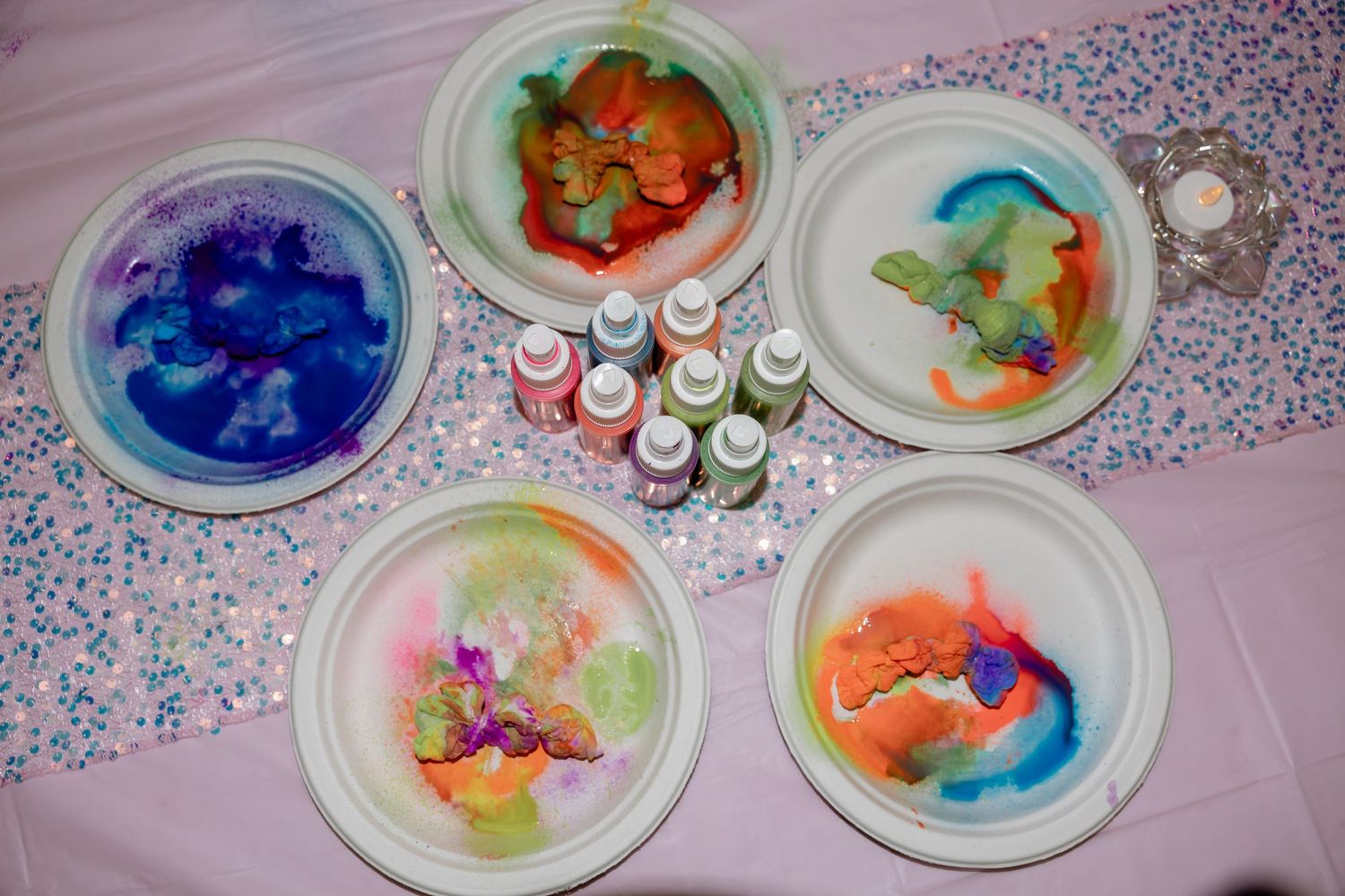 Colorful milk and food coloring science experiment on white paper plates with paint bottles on a pink glitter table.