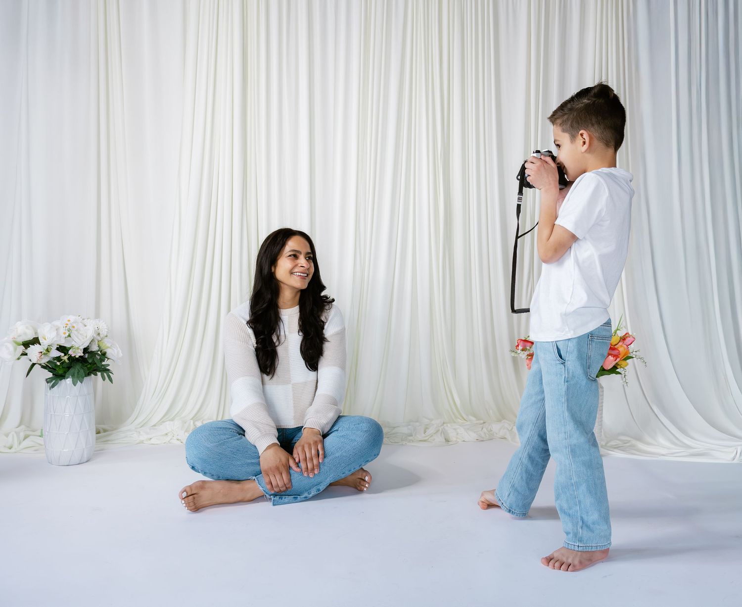 A young boy photographs a smiling woman sitting cross-legged on a white studio floor with floral backdrop.