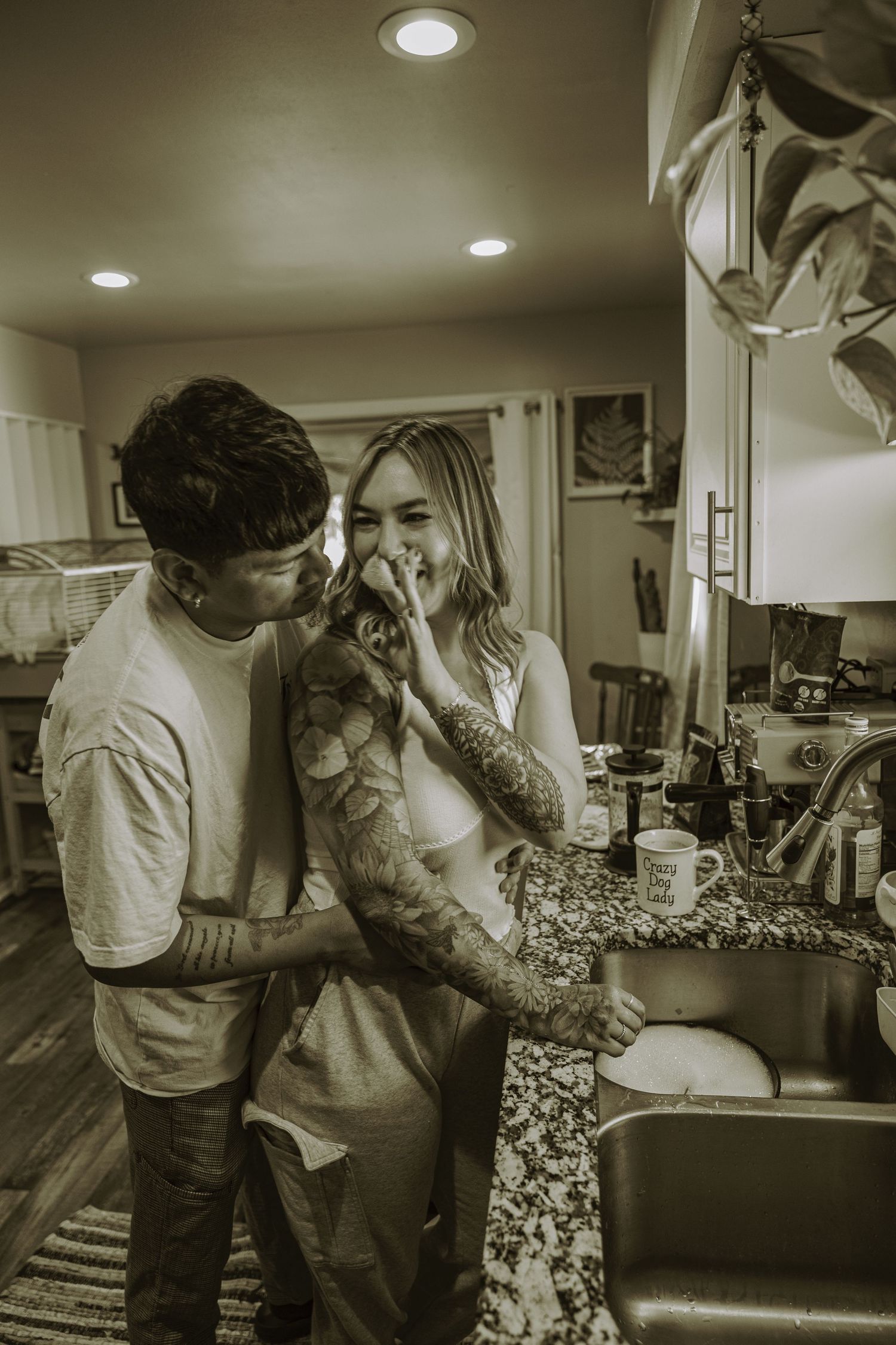 Couple sharing a tender moment in kitchen, man with tattoos embracing woman by the sink, sepia tone.