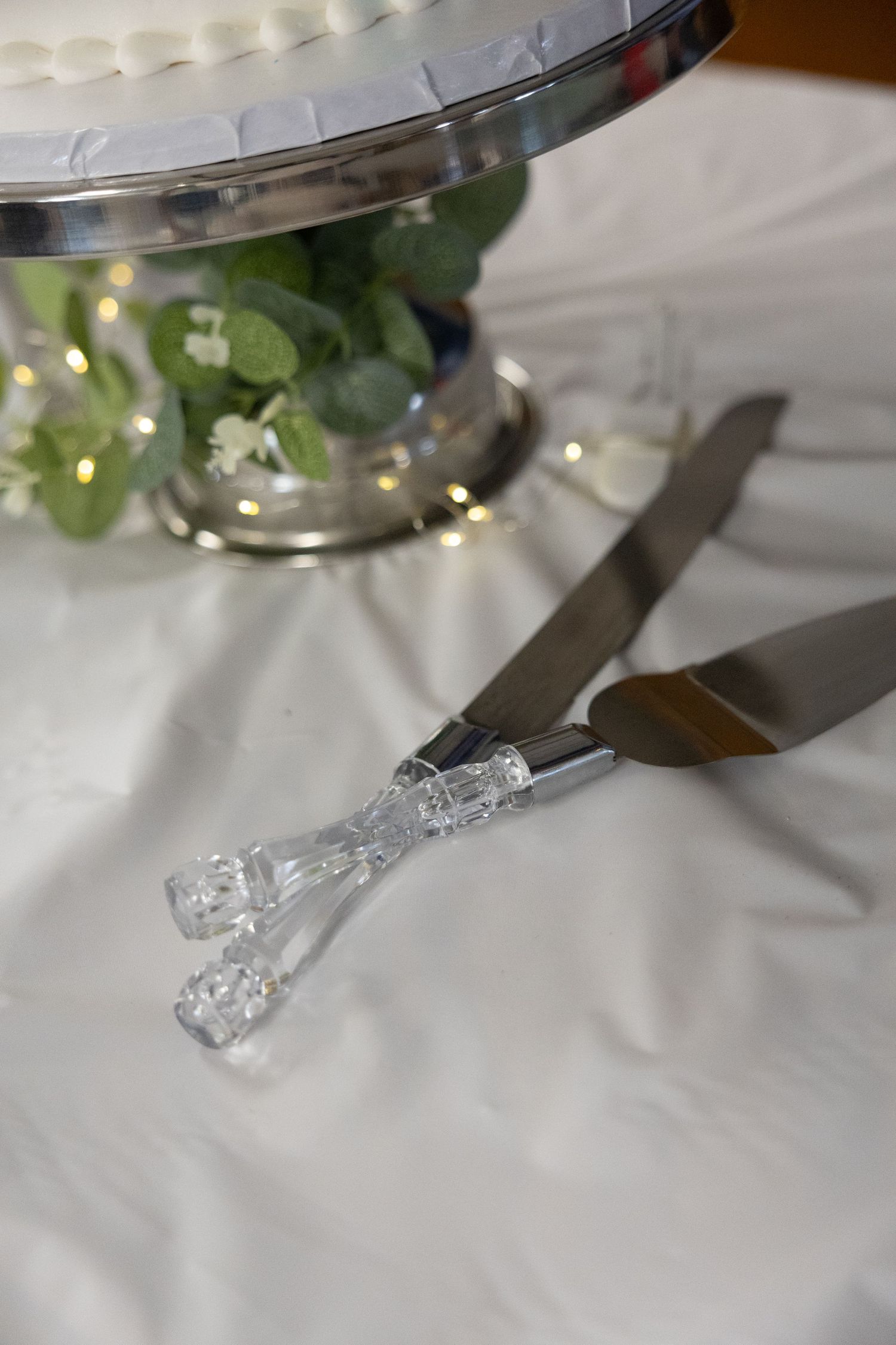Crystal-handled wedding cake knife and server set on white tablecloth beside silver cake stand with floral decor.