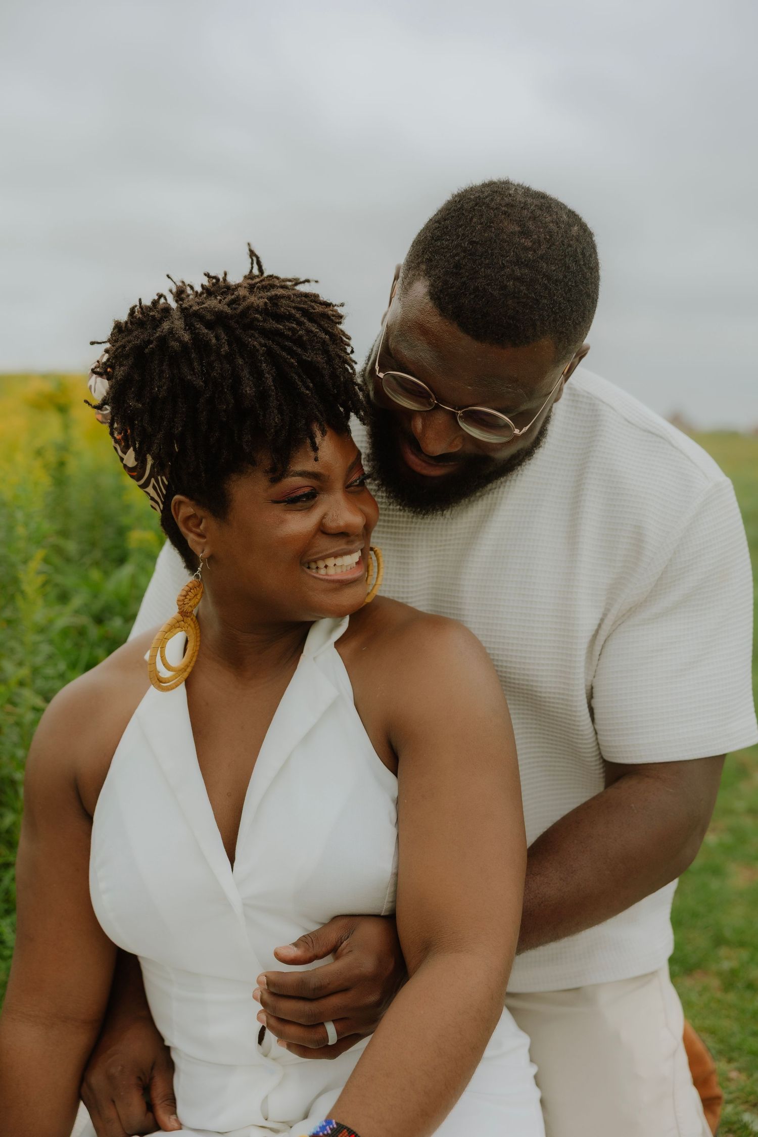 A joyful Black couple embraces in a sunflower field, wearing white outfits, as the man nuzzles the smiling woman.