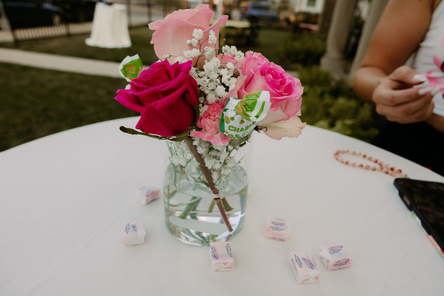 Pink and red roses with baby's breath in a glass vase on a white table with pastel candies at an outdoor event.