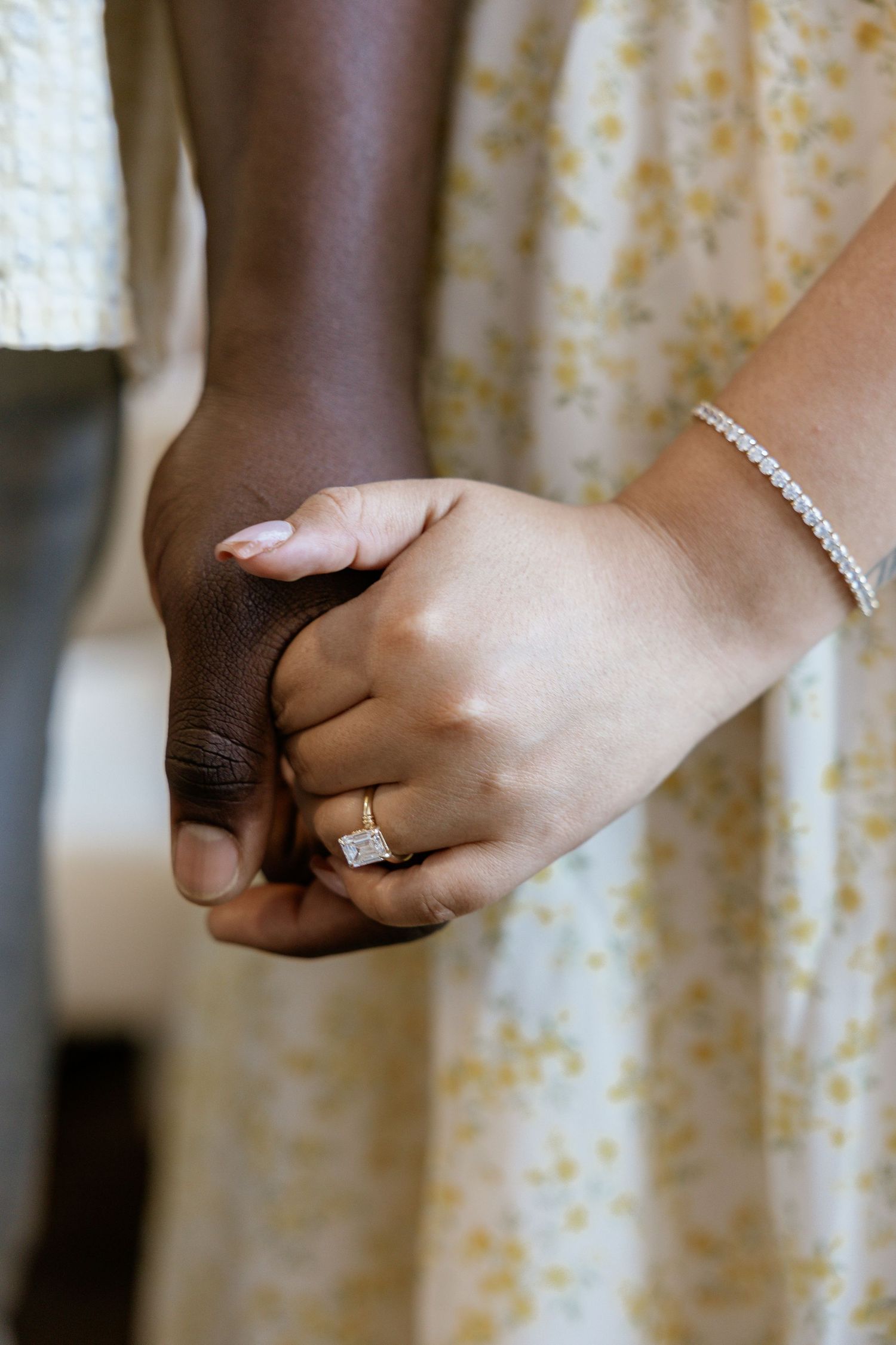 Interracial couple holding hands, woman wearing diamond engagement ring and tennis bracelet, floral dress.