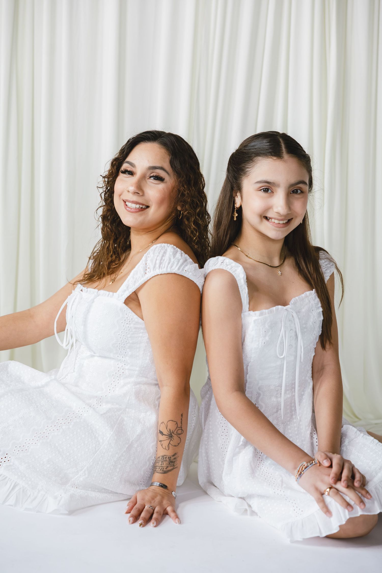 Two women in matching white dresses sit back-to-back, smiling, one with curly hair and a floral tattoo.