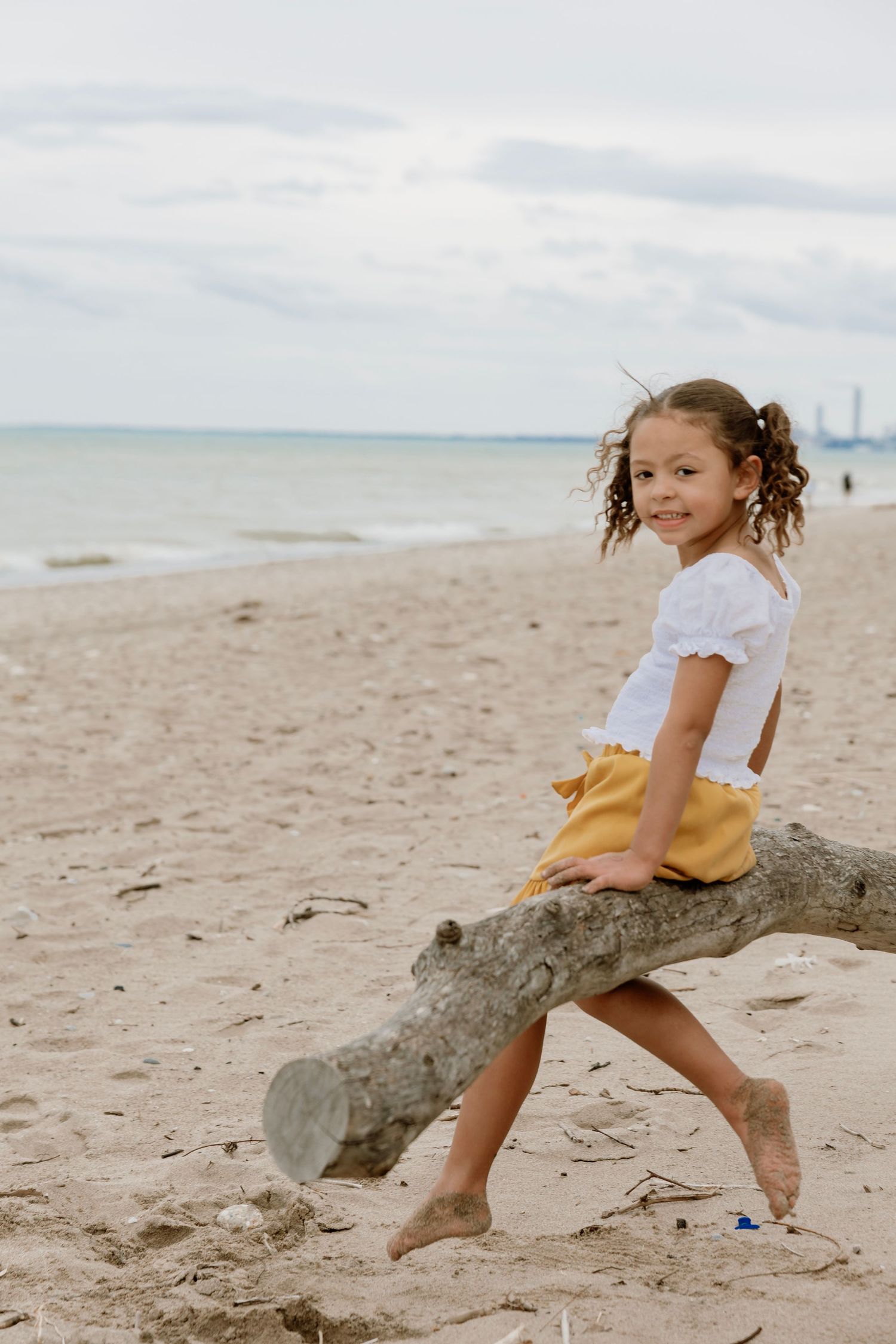 Young girl with curly hair sits on driftwood log at sandy beach, wearing white top and yellow shorts.