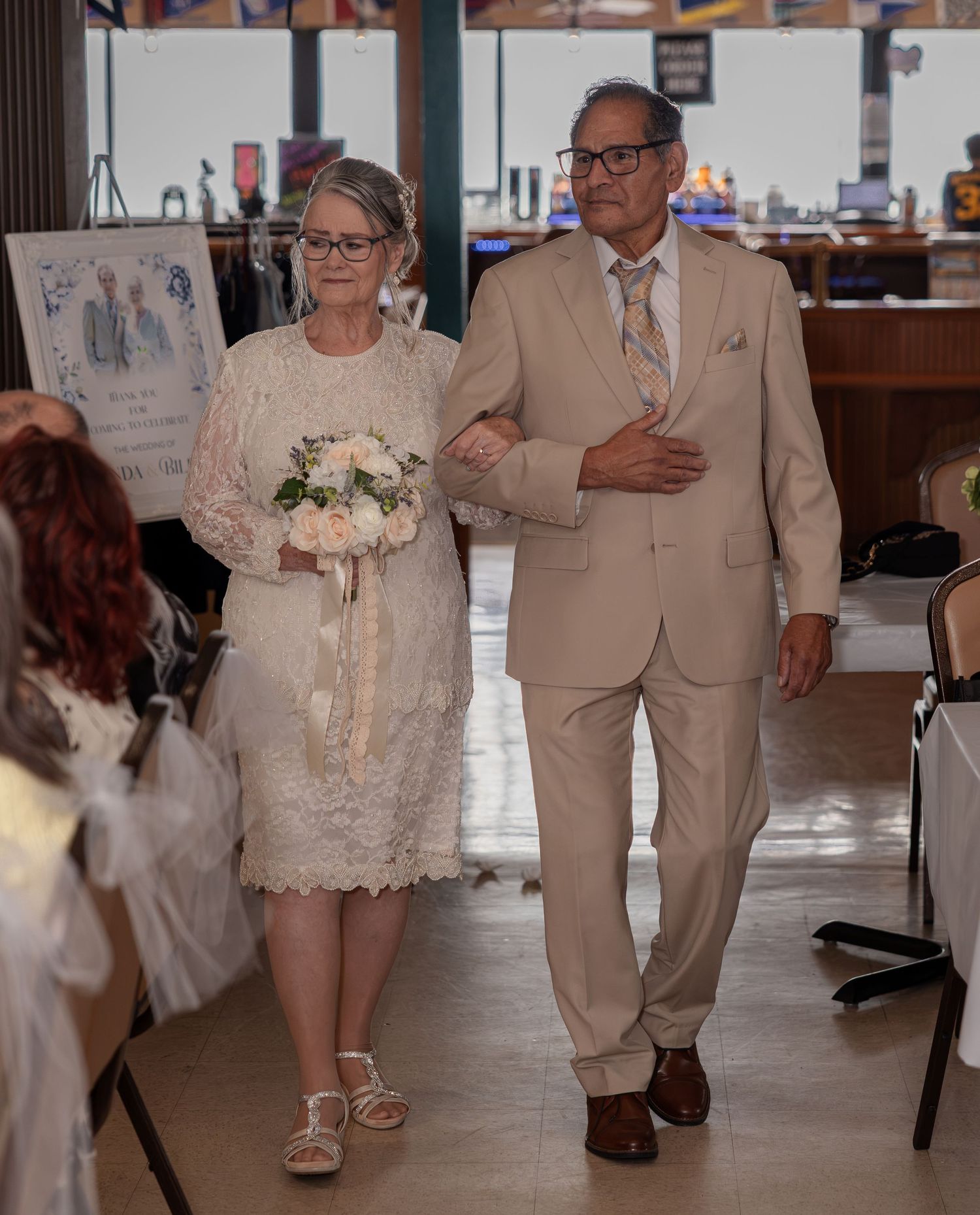 Elderly couple walking down the aisle at a wedding ceremony, woman in lace dress holding bouquet, man in beige suit.