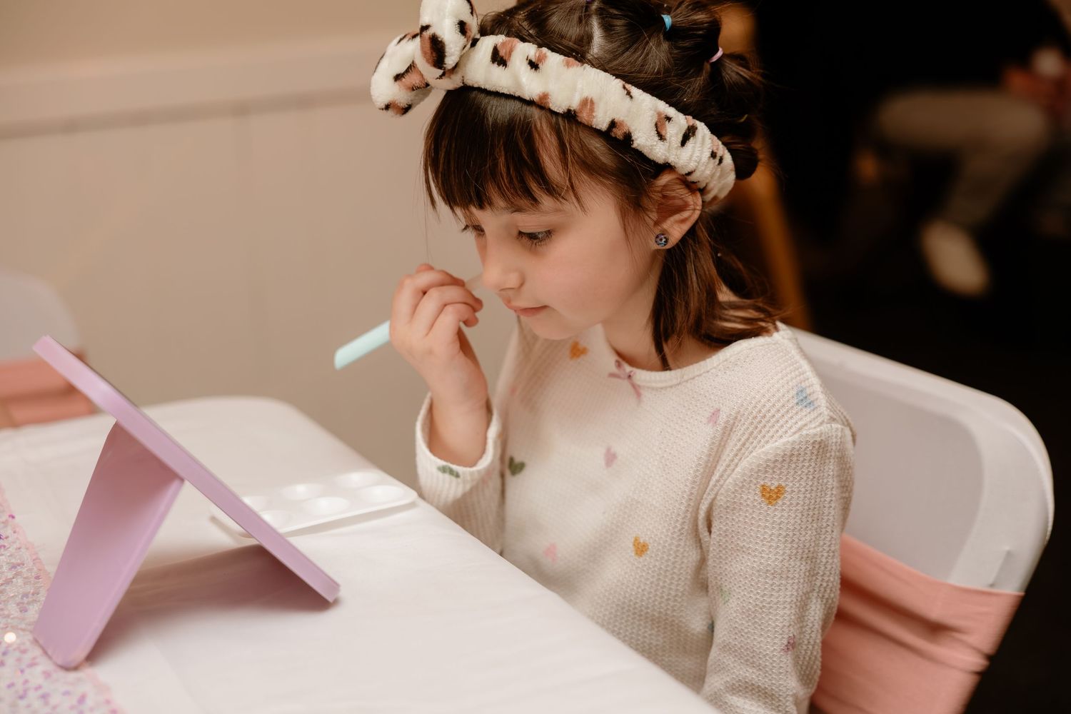 Young girl wearing a headband applies makeup with a brush while looking into a small mirror at a table.