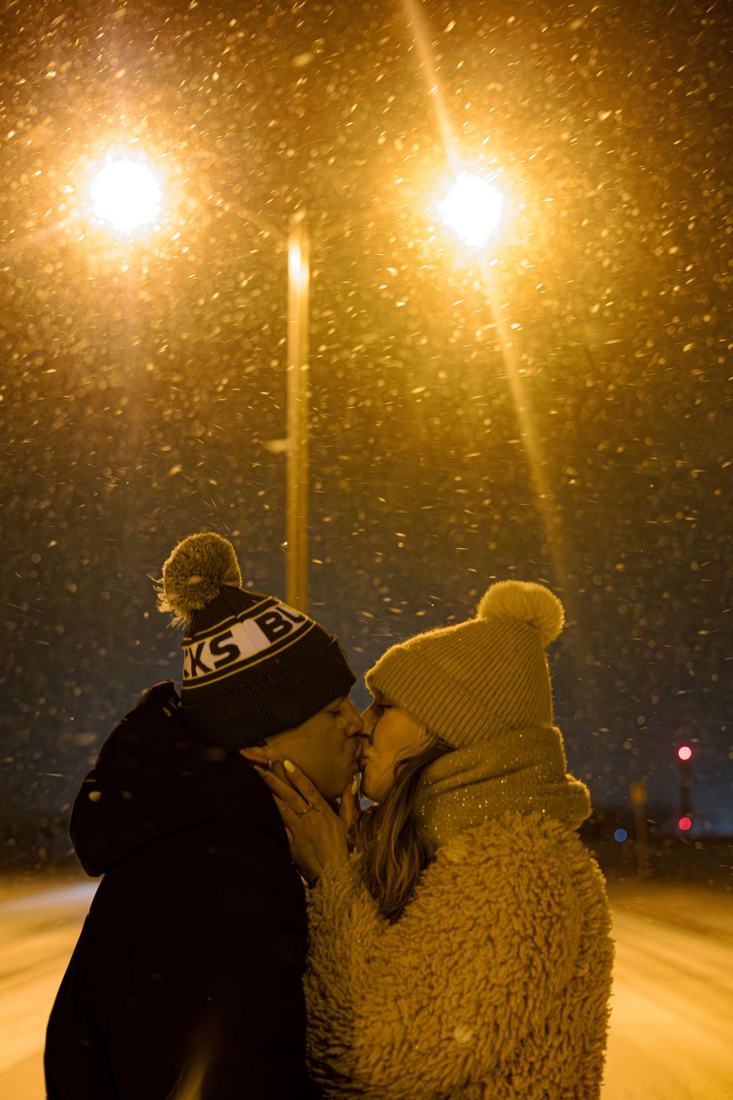 Couple kissing in snowfall under glowing street lights at night, wearing winter hats and cozy coats.