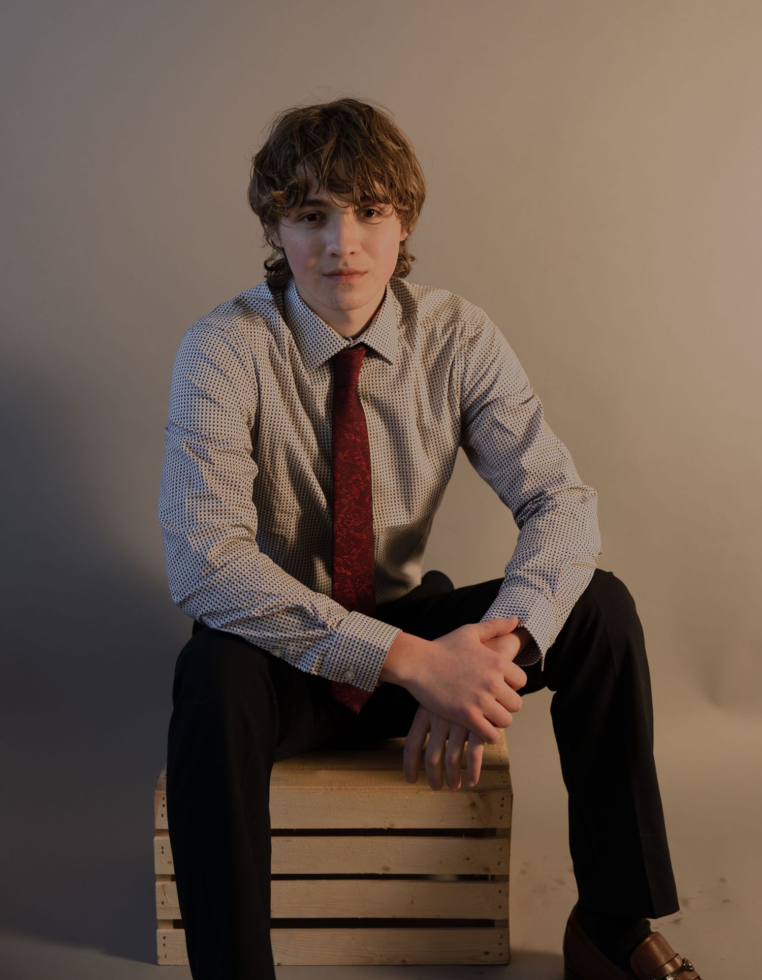 Young man in dress shirt and red tie sitting on wooden crate in professional portrait photo.