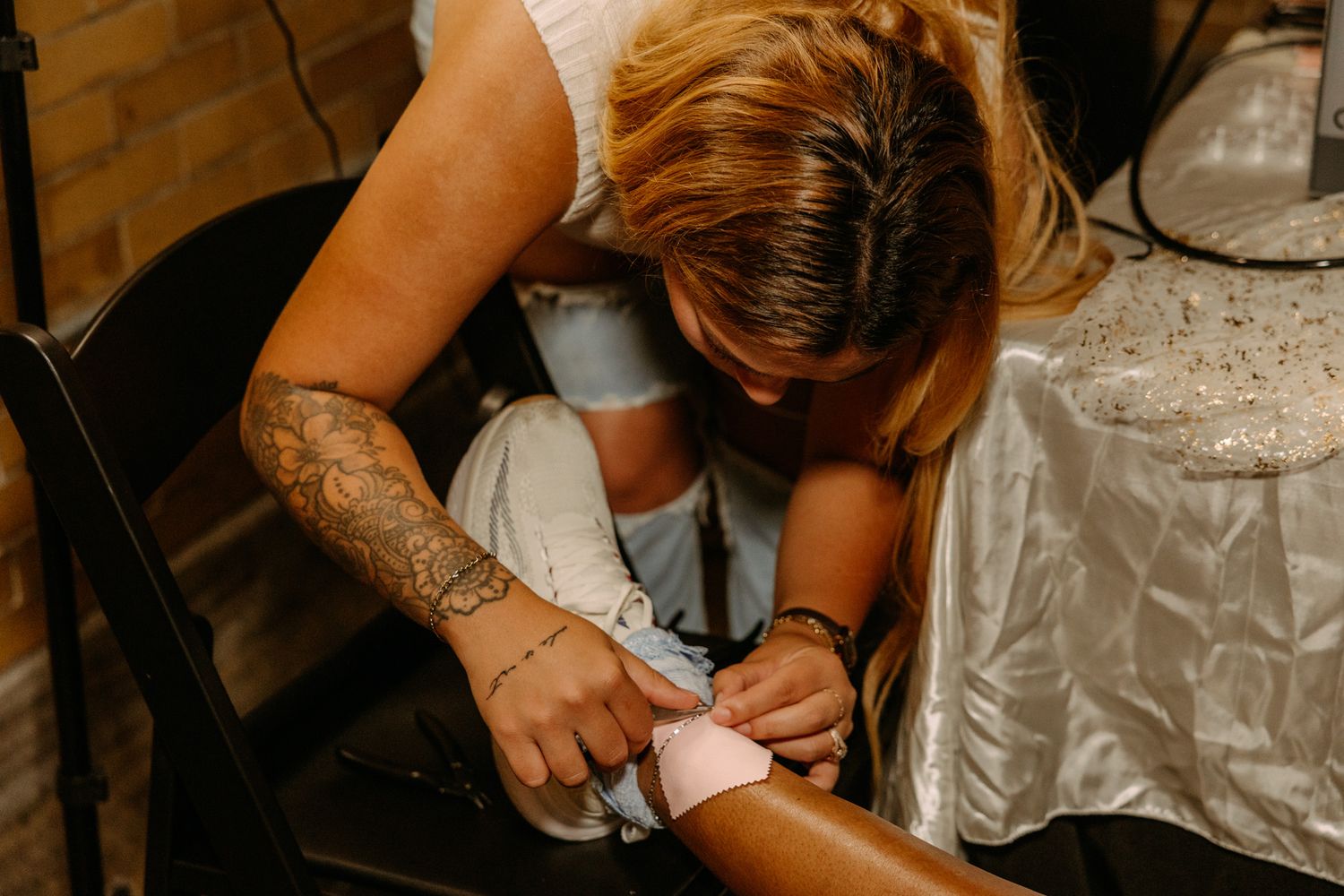 Tattooed bridesmaid helping bride with wedding shoe or accessory, bending down attentively in elegant white dress.