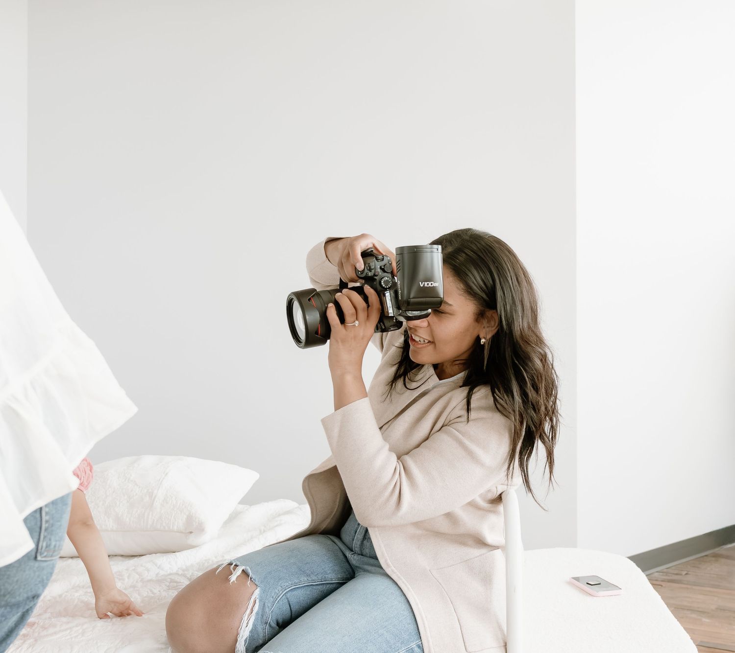 Woman photographer taking photos with DSLR camera while sitting on white bed in bright minimalist room.